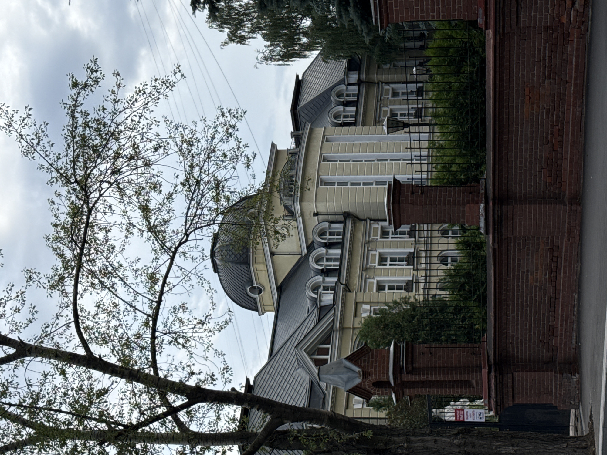 The image depicts a large, historic building in Moscow, Russia, likely a former noble estate or government building. The building is cream-colored with a dark, domed roof and multiple pitched roofs with dormer windows. It features a classical architectural style with columns and ornate detailing around the windows. A tall brick wall with a decorative iron fence encloses the property, separating it from the street. Trees and foliage surround the building, and power lines are visible in the sky. The overall impression is one of grandeur and historical significance, typical of Moscow's older districts. The area appears to be a quiet, well-maintained street with a sense of history.
