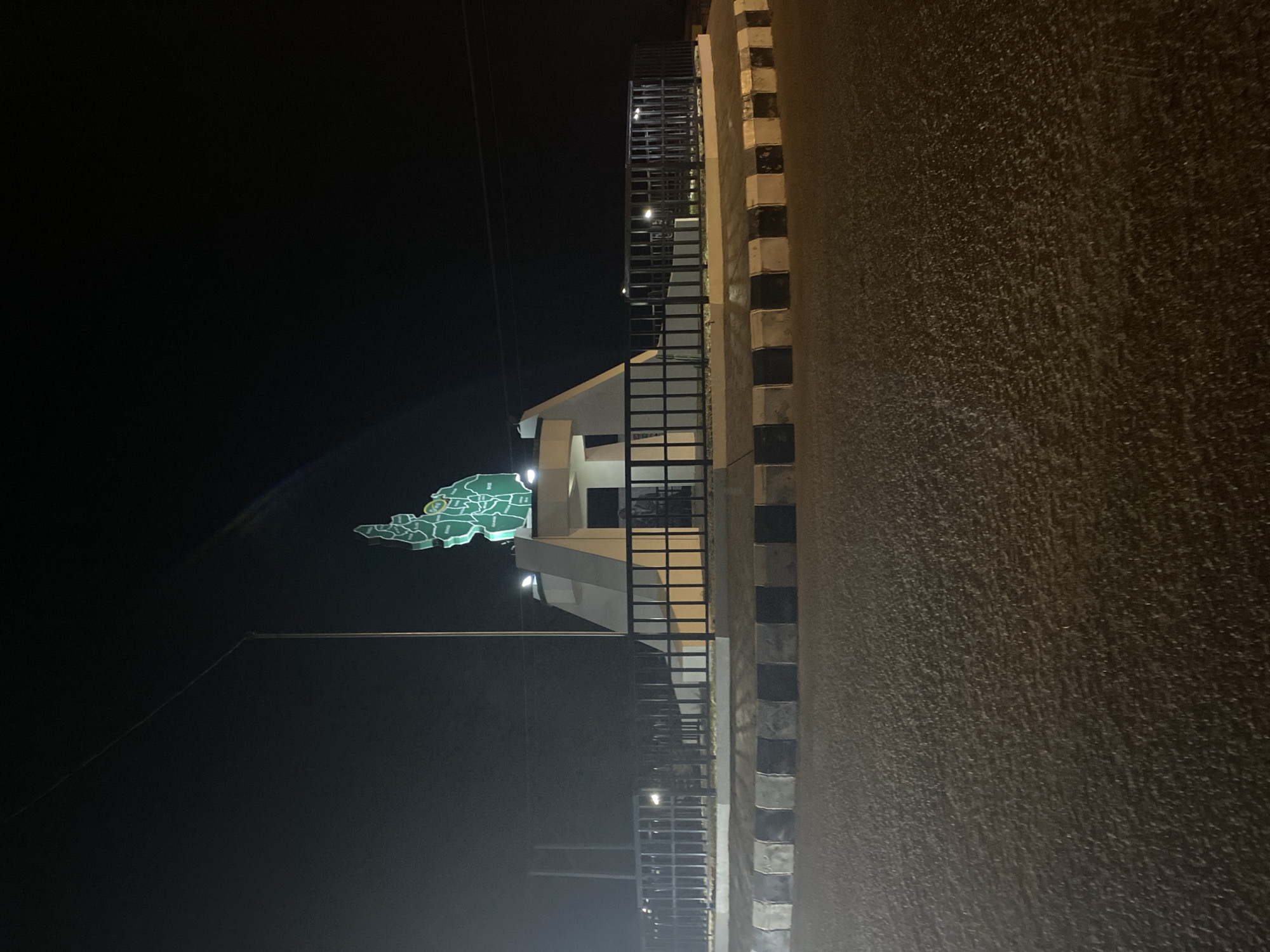 A night-time photograph captures a static scene in Bukuru, Nigeria, dominated by an illuminated green map-like structure and adjacent architectural elements. The image is oriented vertically but depicts a horizontal landscape.
On the left side, a large, bright green map is suspended against the dark sky. This map outlines a geographical area, likely a state or region, showing internal divisions in lighter green tones and circular markers. One prominent circular marker, slightly right of the map's center, appears to contain unreadable text, possibly highlighting a significant location.
To the right of the map stands a building or structure with a light-colored facade, illuminated by internal or external lights. A long, dark metal fence or railing runs along the side of this structure, extending into the foreground. The foreground, occupying the right portion of the image, consists of a textured, dark ground surface, likely a road or paved area, softly lit. Along the far right edge of the frame, a curb or low wall displays a distinct pattern of alternating black and light-colored blocks.
The time of day is clearly night, evidenced by the deep black sky and the sole reliance on artificial lighting to illuminate the map and structure. There are no people visible, and no specific activity is taking place. The scene appears to showcase a public landmark or informational display. Due to the distance and resolution, specific text on the map is not discernible beyond its presence.