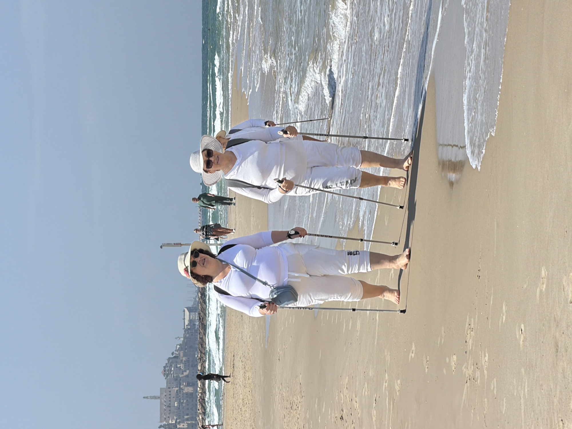 Two elderly women are walking on a beach in Tel Aviv, Israel, each using Nordic walking poles. They are dressed in white clothing and hats. A distant view of the city's skyline, including buildings and a tower, is visible in the background. Others are walking along the beach further back. The ocean is to their right, with waves washing ashore. The sand is light in color, and the sky is clear.