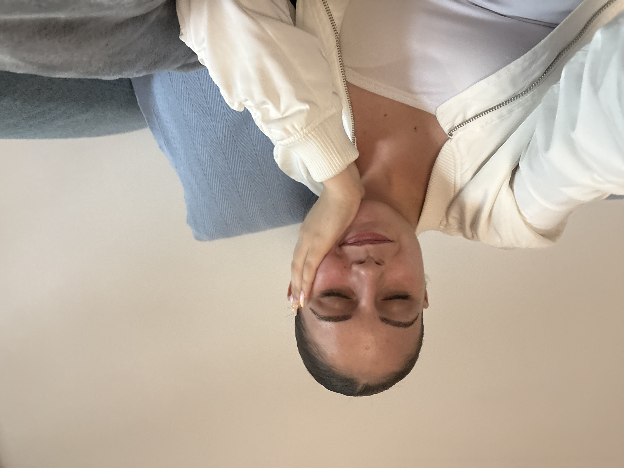 A woman is reclining, appearing to be either lying down or leaning back at an extreme angle relative to the camera. Her eyes are closed. She is wearing a white top and a white zip-up jacket. Her hand is resting on her cheek. A blue pillow and a gray blanket are visible behind her. The background is a plain, light beige or cream-colored surface. There is no visible evidence to support the determination of a specific location such as the city of Burgas, Bulgaria.