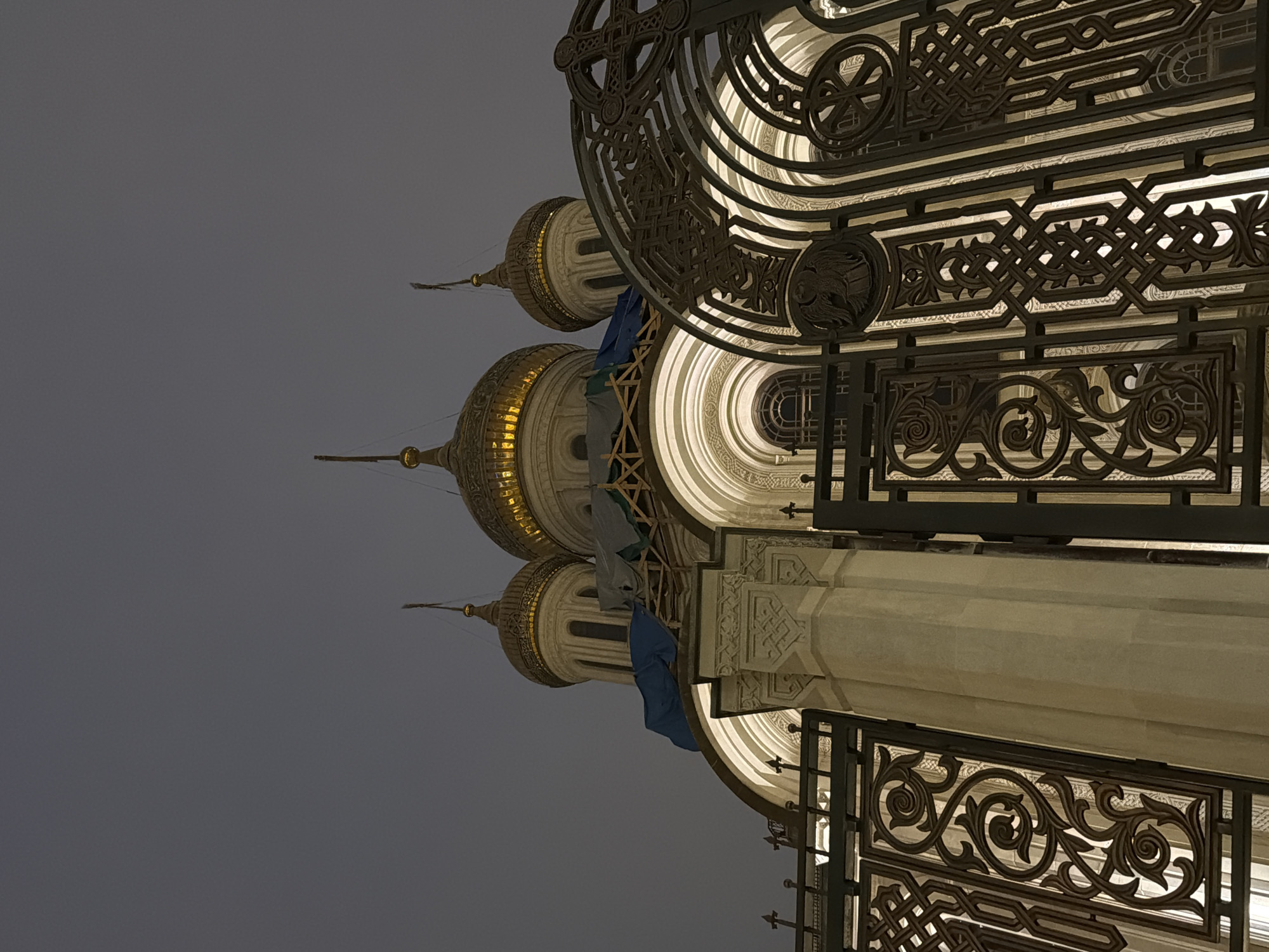 The image shows an upward-angled, nighttime view of a building in Moscow, Russia, featuring ornate golden domes and intricate wrought-iron detailing. The domes, traditional in Russian Orthodox architecture, have a reflective golden surface and are topped with spires. Scaffolding with blue and white covers is visible near one of the domes, suggesting ongoing restoration or maintenance. The wrought-iron work displays a pattern of concentric circles, geometric shapes, and floral motifs, and is illuminated from within. The background sky is a solid, neutral gray. A stone pillar or wall segment is also visible, decorated with architectural patterns. The structure appears to be a historical building, likely a cathedral or significant cultural landmark in Moscow.