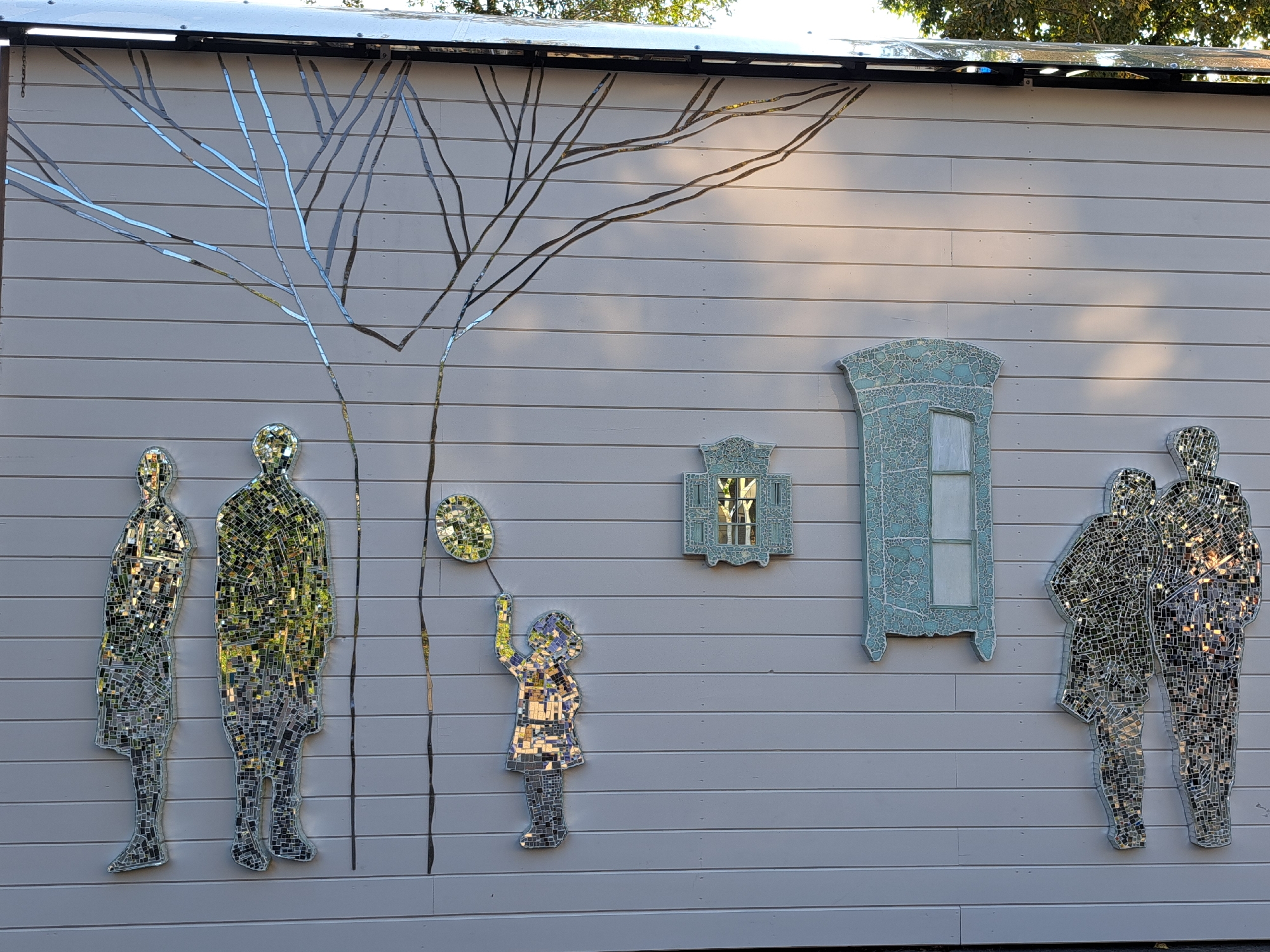 This image depicts an outdoor wall mural made of mosaic tiles on a light-colored, horizontally paneled wall. The mural features several elements:
* **Tree Branches:** Stylized, leafless tree branches are depicted across the top portion of the wall, constructed from thin, reflective mosaic pieces that catch the light.
* **Human Figures:**
* On the left, two full-body figures are rendered in mosaic, appearing as silhouettes filled with reflective tiles.
* In the center, a child figure, also made of mosaic, is shown holding a balloon. The child is depicted in a dress and leggings.
* On the right, another pair of full-body figures, similarly constructed from mosaic, are shown standing close together.
* **Architectural Elements:** Two decorative, window-like structures with blue, crackled mosaic tiles are mounted on the wall. The larger one on the right resembles a closed window with panes, while the smaller one on the left has a more ornate, decorative frame.
* **Balloon:** A single, circular mosaic element, representing a balloon, is held by the child.
* **Environment:** The wall is set against an outdoor background with hints of trees and sunlight filtering through, suggesting an exterior setting. The overall impression is a whimsical and artistic display on a building's side.
This artwork is located at the Irkutsk Regional Art Museum in Irkutsk, Russia.