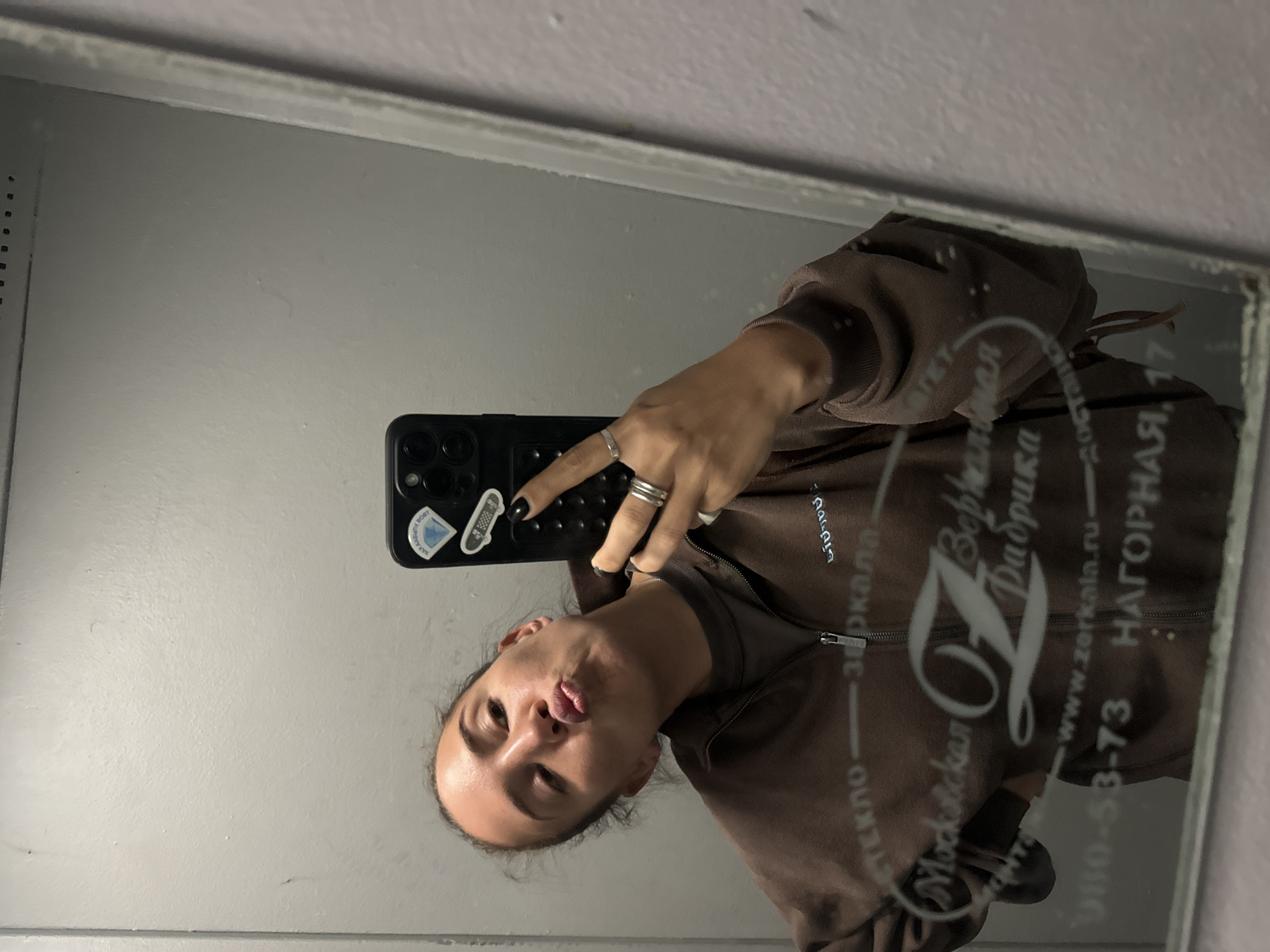 A person takes a selfie in a mirror. The person has dark hair pulled back, a brown zip-up sweatshirt, and several rings on their fingers. They are making a kissing face. The phone has stickers on the back. Reflected in the mirror is the text "Московская зеркала Верфь". It reads, "зеркала зстекло" and "НАГОРНАЯ, 17". The phone number "+7 495 980-53-73" and "www.zerkala.ru" are also visible. This indicates the location may be in or near Khimki, Russia, specifically on Nagornaya Street. The surrounding environment appears to be a plain gray wall.