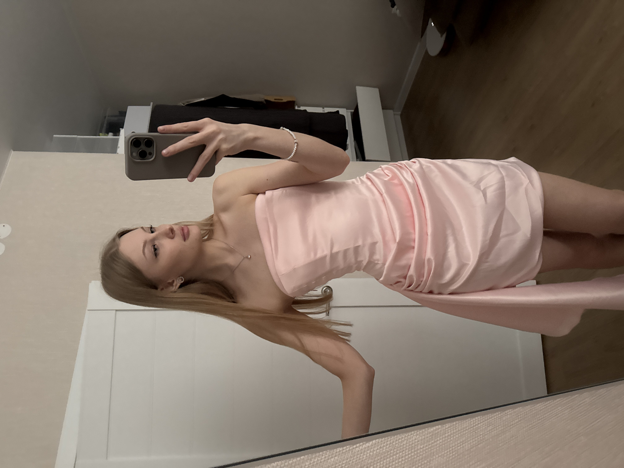 A young woman with long blonde hair is taking a selfie in a mirror. She is wearing a short, pink dress and jewelry. The background includes a white wardrobe, a bed or sofa with dark upholstery, and a wood-paneled wall. The location is likely indoors. There is no definitive evidence indicating the city of Perm, Russia.