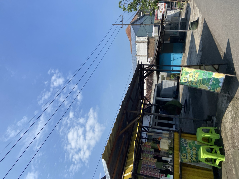 A street scene in Mojosari, Indonesia, located at coordinates -7.503 latitude and 112.54 longitude. The scene is a street with a yellow and black food stand on the left side of the frame. The food stand has a sign that reads "Es Jeruk Peras" which translates to "Orange Juice." The sign has the price "Rp 5000" written on it. There are two green plastic stools in front of the stand. There are various items on the shelves of the stand, including plastic bags of food, a small metal blender, and a variety of other items. There is a large, round, green object hanging from the ceiling of the stand. On the right side of the frame, there are two small buildings, one blue and one gray. There is a large, brown trash can and a small, white sign with the text "Bag Low" on it in the distance. The buildings are made of concrete and brick. The scene is under a clear, blue sky with some white clouds. There are power lines running across the scene.