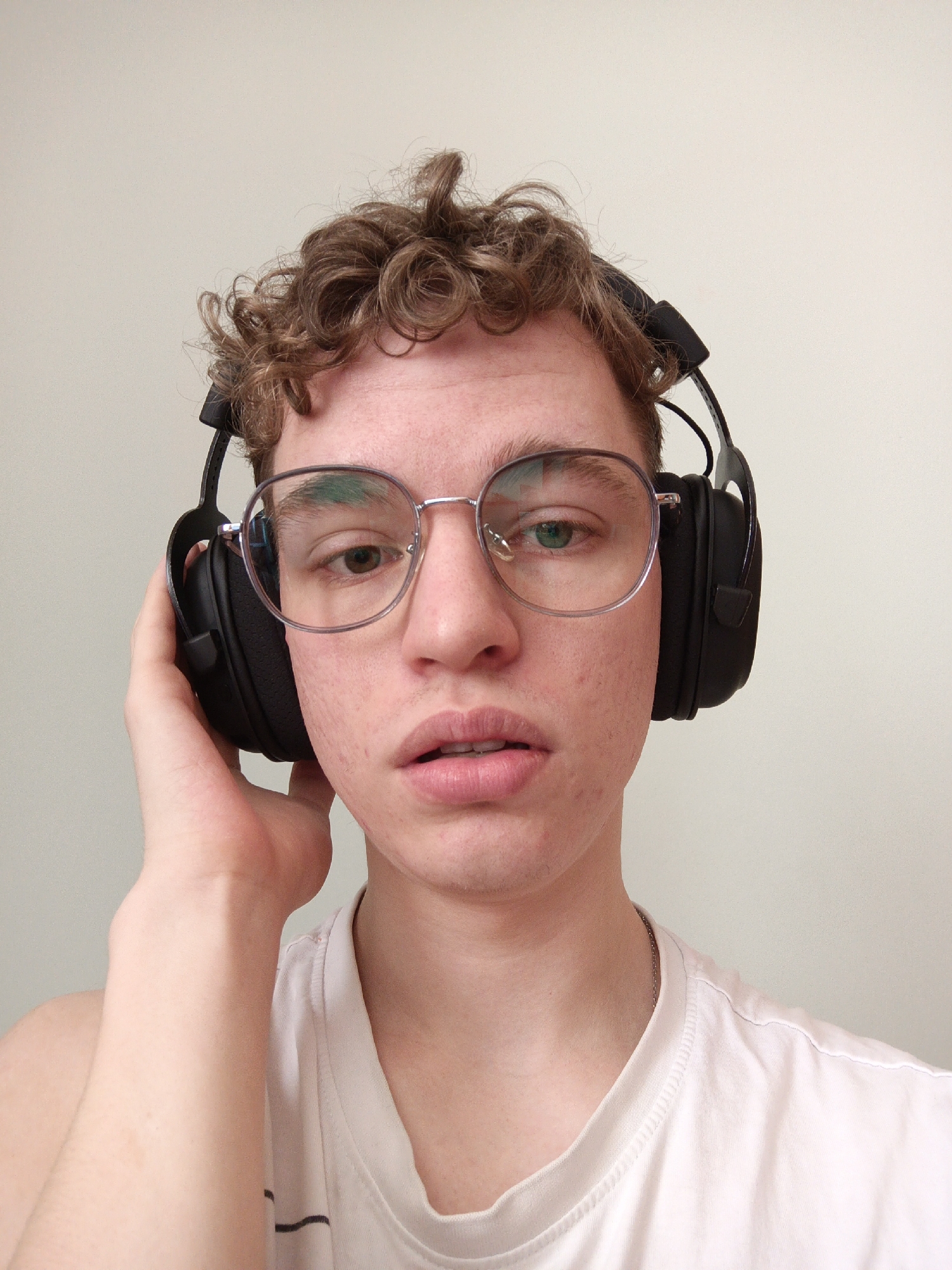 A young man with curly brown hair and silver-rimmed glasses is wearing black over-ear headphones. He is holding the right earcup with his right hand. He is wearing a white t-shirt. The background is a plain off-white wall. The image provides no information about the city of Odintsovo, Russia.