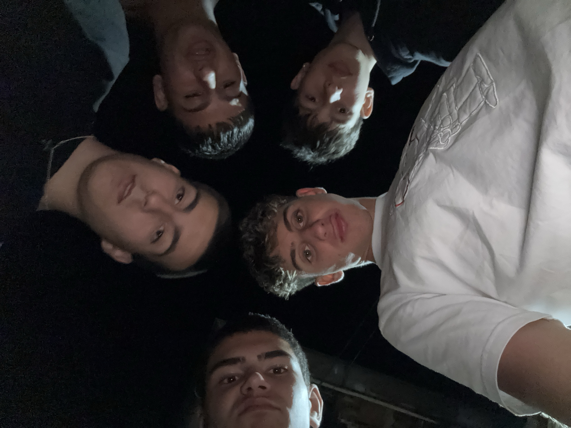 This is a medium shot, low-angle, portrait-style photograph taken at night, looking up at five young males. They are gathered in a huddle, with their faces and torsos filling the frame from the perspective of someone lying on the ground or very low. The individuals are all looking down at the camera. The setting appears to be outdoors, with a dark background that suggests a lack of ambient light, possibly indicating a street or alleyway. The lighting is artificial and harsh, creating strong highlights and shadows on their faces and clothing. The individuals are casually dressed, with one in the foreground wearing a white t-shirt with an embroidered graphic. The overall impression is of a candid, possibly impromptu, gathering.
