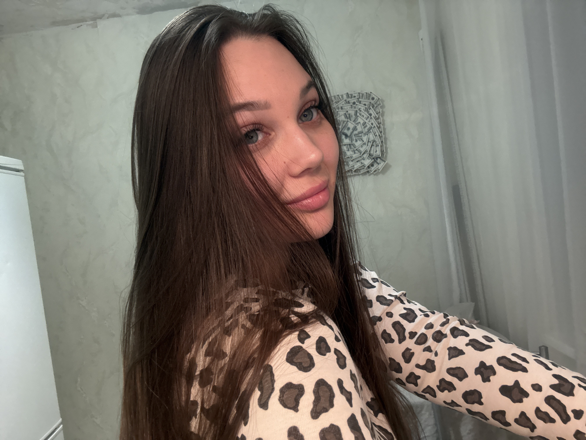The image presents a portrait of a young woman taken indoors, likely in Chaikovsky, Russia. She has long, straight dark brown hair, blue eyes, and full, pink lips, with a soft, gentle smile directed towards the viewer. She is wearing a long-sleeved top featuring a light-colored background with a dark leopard print pattern.
The background indicates an interior space, possibly a room in a home. To the left, a portion of a white appliance, potentially a refrigerator or cabinet, is visible. The walls are a light grey with a textured finish. A distinctive piece of wall art, appearing to be made from folded or crumpled dollar bills arranged in a circular or fan-like design, hangs on the wall to the right behind her. Further right, a light-colored curtain or fabric is partially visible. The lighting is even, suggesting an indoor setting. The woman's expression conveys a sense of calm and slight allure. No visible text is clearly legible within the image.
