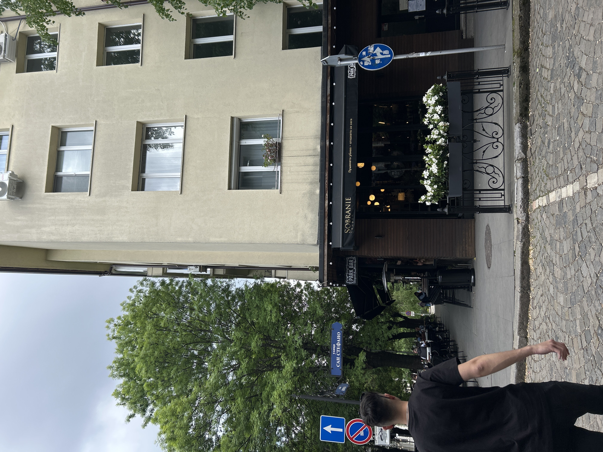 The image shows a street scene, likely in Sofia, Bulgaria, based on the Cyrillic signage. A light-colored building with several windows occupies the left side of the frame. The restaurant "Sobranie" is located on the ground floor of the building. A wrought-iron fence with white flowers is near the restaurant entrance. A blue street sign with Cyrillic script, "улица Сан Стефано" ("ulitsa San Stefano", or San Stefano Street), is visible near the sidewalk. Street signs with pedestrian crosswalk are visible. A man in a black t-shirt is partially visible in the foreground, walking on a cobblestone sidewalk. Several trees obscure the restaurant patio.