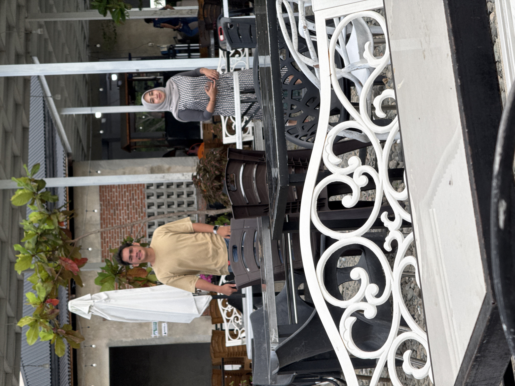 In Cotmalem, Indonesia, a man in a tan t-shirt and a woman in a grey hijab and striped dress stand near each other in an outdoor dining area. The man is looking towards the camera, while the woman is looking down at her hands. Behind them, there are tables and chairs, as well as trees and a building. The overall atmosphere appears casual and relaxed.