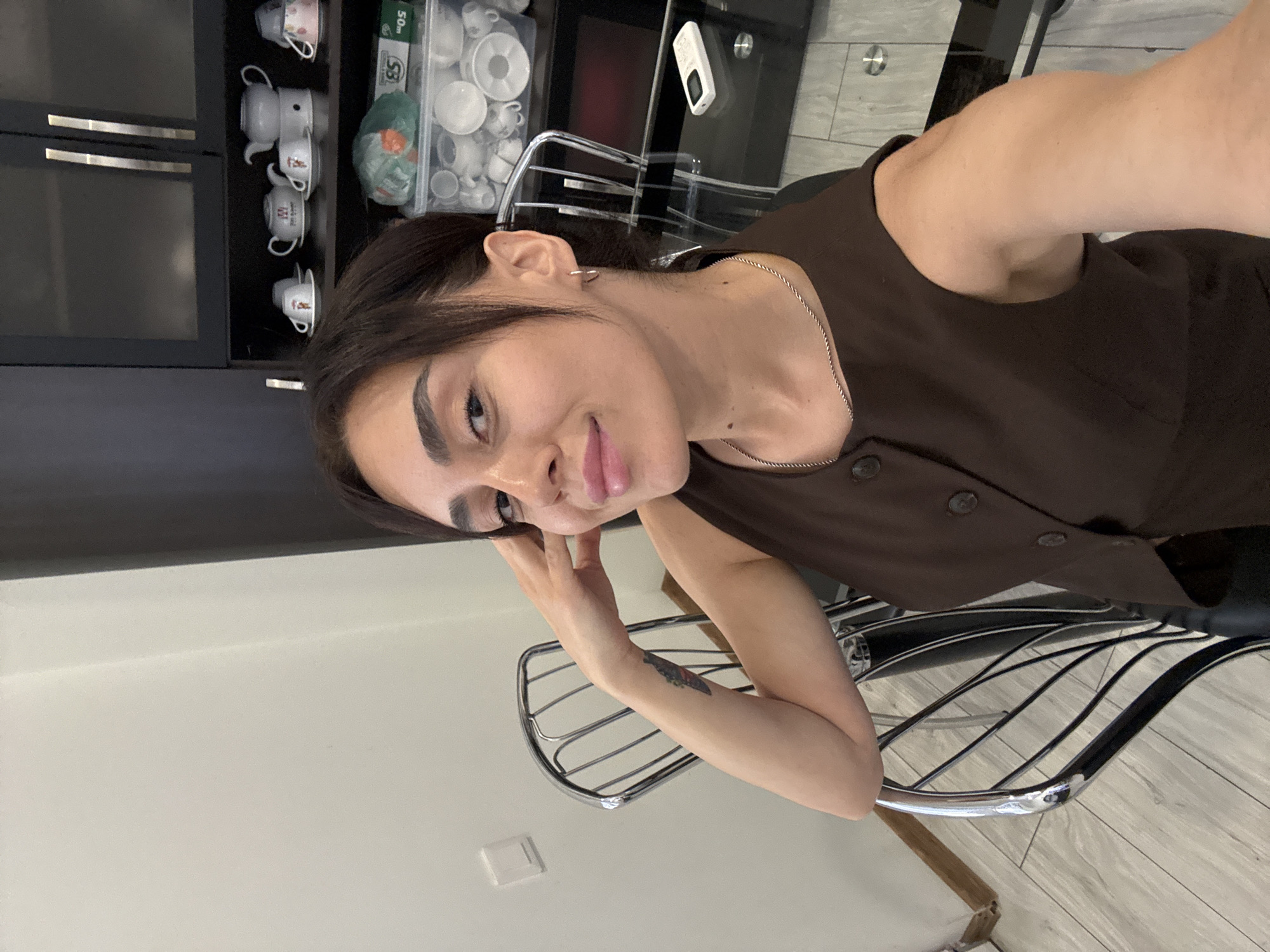 A woman is captured in a selfie, seated indoors at a table. She has dark hair pulled back, light skin, and is wearing a brown sleeveless vest and a silver necklace. Her left arm is bent, resting on the back of a metal chair. The background reveals a wall, a portion of a cabinet with teacups and saucers, and a transparent table with a remote control. The setting appears to be a domestic interior, possibly a dining area or kitchen. There is no visible indication that this location is in Tbilisi, Georgia.