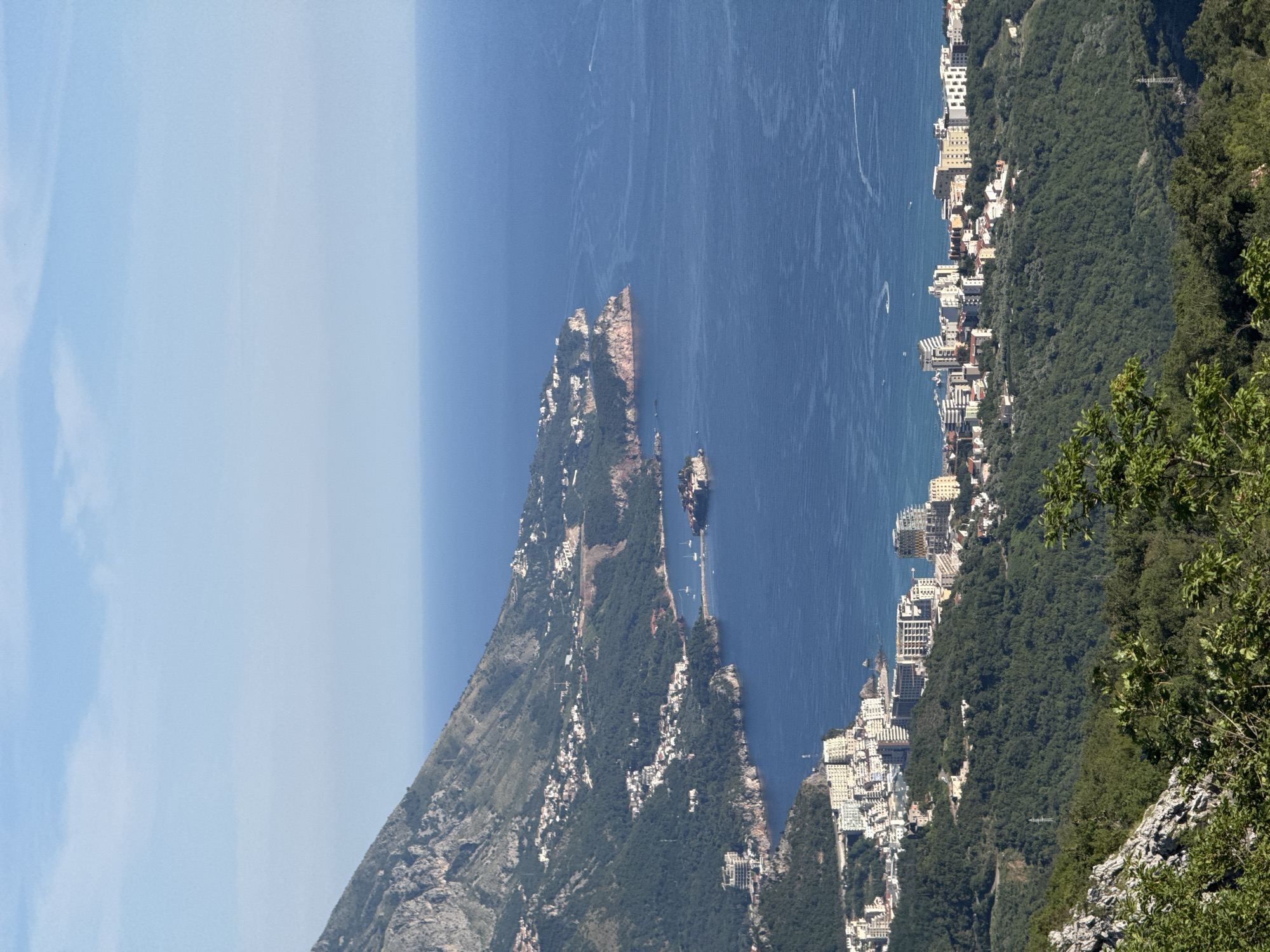 This is a high-angle, wide shot of a coastal city with mountains in the background. The city is built on hillsides overlooking the blue ocean. Numerous buildings, ranging from smaller homes to multi-story hotels, are visible along the coastline and extending up the hills. Several boats are scattered on the water. The sea is calm with some gentle ripples. The sky is a clear blue with a few wispy clouds. The foreground shows the rocky, green slopes of a mountain, with some foliage obscuring the view. A small island with buildings is connected to the mainland by a causeway in the middle distance.