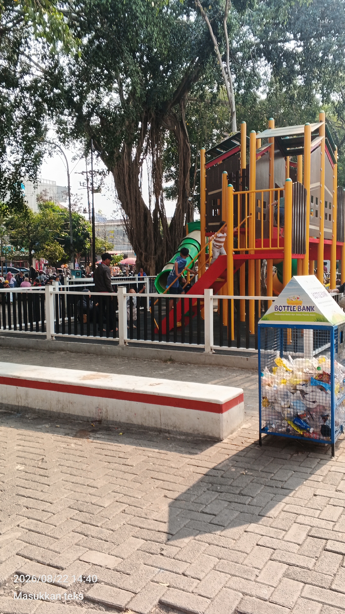 This image captures a bright, sunny day at a public park in Malang, Indonesia. In the foreground, a light-colored bench with a red stripe runs horizontally across the frame, positioned on a brick-paved walkway. To the right of the bench, a blue mesh bin is filled with plastic bottles and other recyclable materials, topped with a white sign that reads "BOTTLE BANK" in Indonesian.
Behind the bench, a black metal fence encloses a children's playground. The playground features a large, multi-colored structure with yellow poles, a red slide, and a green tunnel slide. Several children are visible playing on the structure, including one boy in a blue shirt on the stairs and another in a striped shirt on the red slide.
In the background, a large, ancient banyan tree with its characteristic aerial roots dominates the left side of the image. Beyond the tree and playground, a bustling scene unfolds with a crowd of people, some appearing to be shoppers or visitors. Street lamps and distant buildings are also visible. The overall impression is a lively, family-friendly outdoor space in an urban setting.
