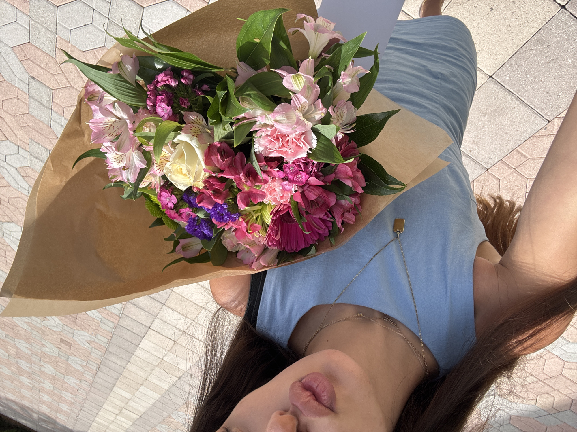 In Boca Raton, United States of America, an outdoor, high-angle selfie depicts a female individual holding a large, diverse bouquet of flowers. The individual's face is partially visible, showing her chin, lips puckered in a kiss gesture, and the lower part of her nose. Her dark brown hair cascades over her shoulders. She wears a light blue sleeveless dress and two thin gold necklaces, one with a small rectangular pendant. The bouquet, wrapped in brown kraft paper, features a variety of blooms including pink and white carnations, pink and purple alstroemeria, dark pink/magenta gerbera daisies, light pink/white roses, and small dark purple flowers, interspersed with green foliage. The background consists of a geometrically patterned pavement, composed of light grey, white, and pale reddish-pink hexagonal and rectangular tiles, under bright daylight.
