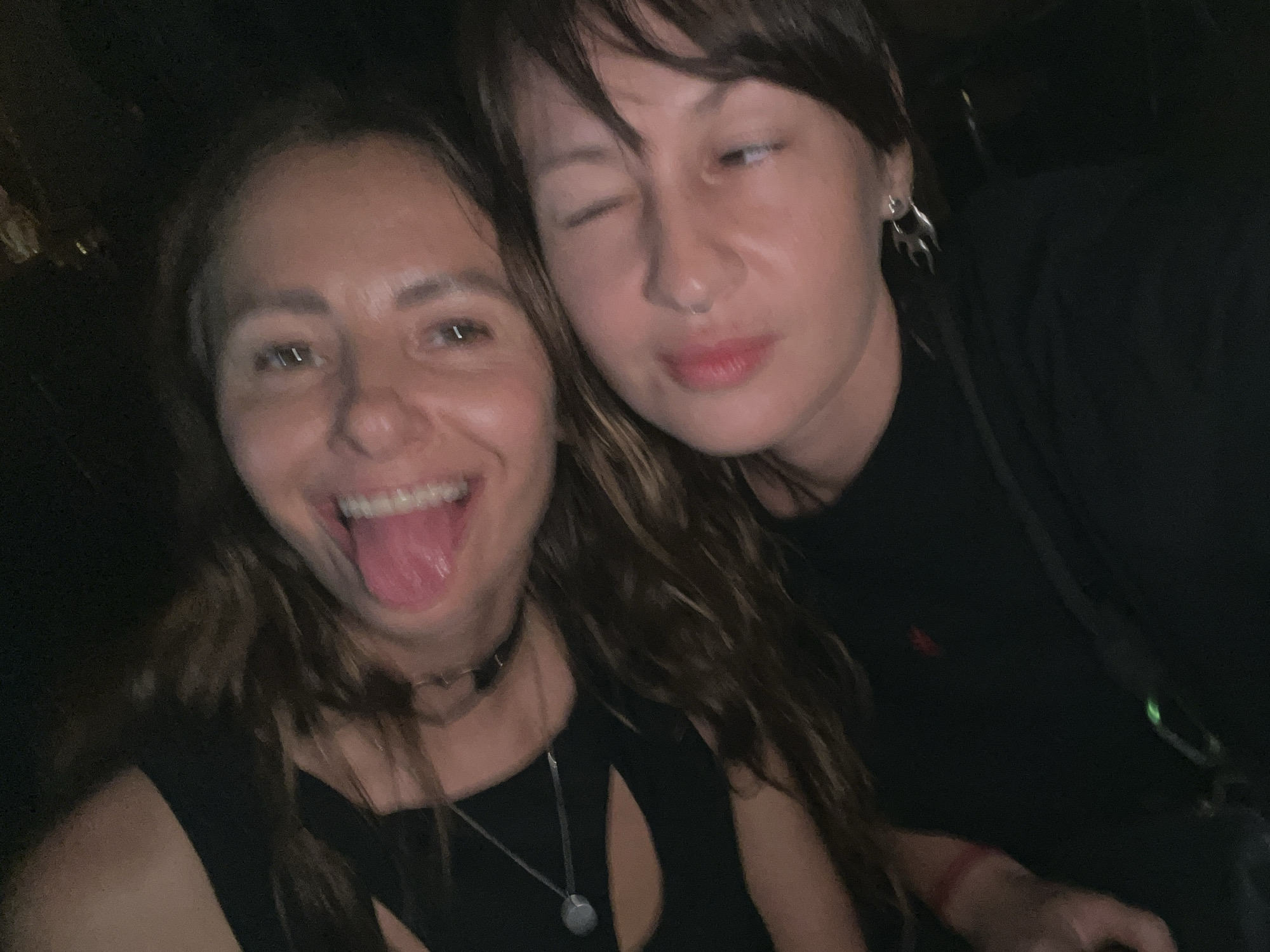 Two women are posing closely together for a photo. The woman on the left has long brown hair, wears a black choker and a black top with cutouts, and has her tongue sticking out. The woman on the right winks, has short dark hair with bangs, and wears a black shirt and silver flame-shaped earrings. The background is dark and indistinct. The location is not discernible from the image, and there is no information provided that indicates the picture was taken in Mengwi, Indonesia.