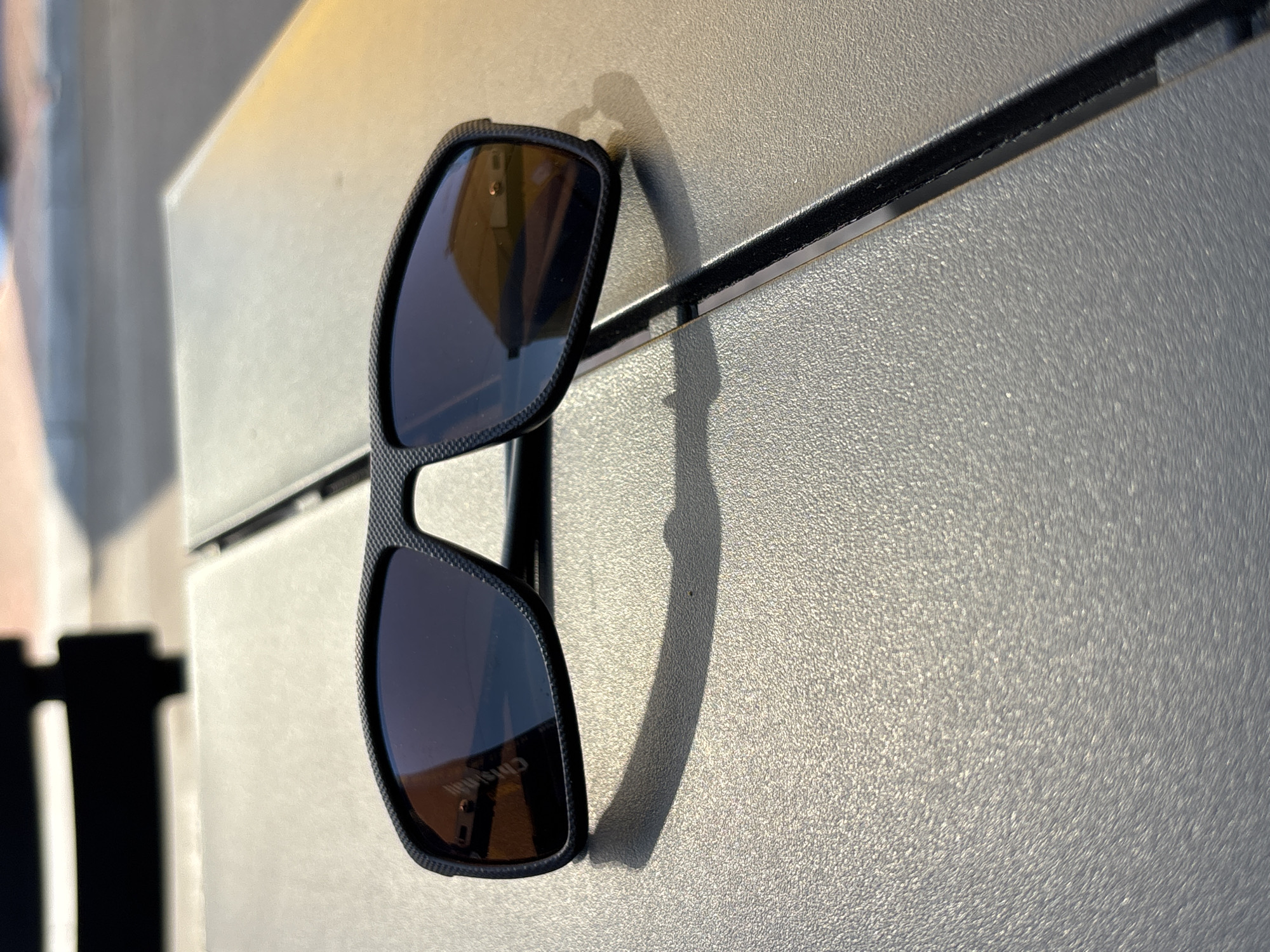 A pair of black, wrap-around sunglasses with dark lenses rests on a dark gray, textured surface. The sunglasses are positioned slightly off-center, with the left lens facing the camera. The surface appears to be an outdoor table, possibly made of plastic or metal, with a visible seam running diagonally across the image from the bottom right to the top left. The background is blurred, showing a suggestion of sunlight and a dark, out-of-focus structure. A faint shadow of the sunglasses is cast on the textured surface. The reflection in the lenses shows an outdoor environment with some yellow and orange hues.