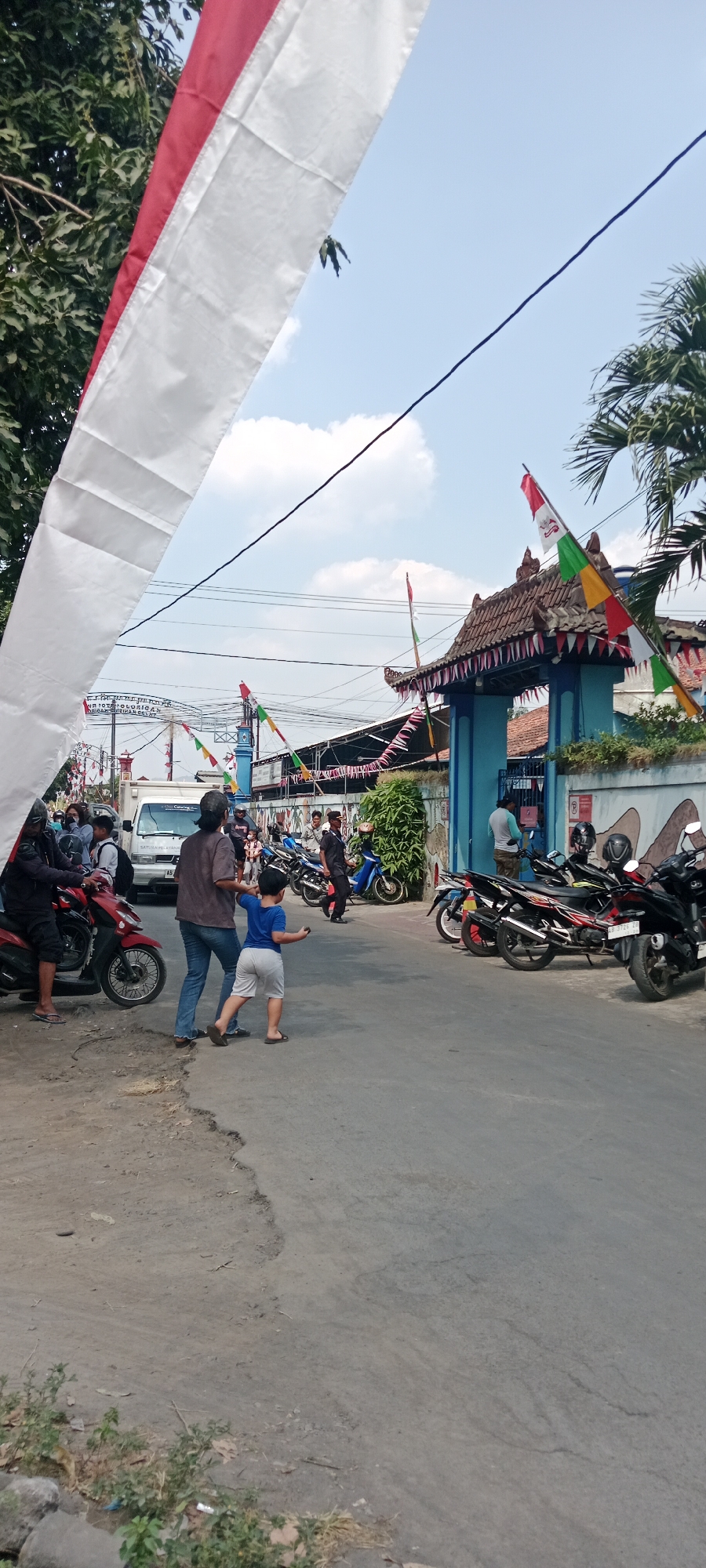 In Purwomartani, Indonesia, a street scene is depicted under a bright, partly cloudy sky. Red and white Indonesian flags and pennants adorn the area, strung across the street and from a building entrance, suggesting a celebration or event. A white van is parked on the left side of the road, with several people standing around it. In the center of the image, a woman in a brown shirt and jeans stands next to a young boy in a blue shirt and gray shorts. The boy is facing away from the camera, with his arms extended as if throwing something. A red motorcycle is parked to the left of the woman and boy.
Further down the street, several motorcycles are parked in front of a blue building with an ornate, tiled roof. More people are gathered near the building entrance, some standing and some appearing to be engaged in conversation. A tall, slender blue structure, possibly a minaret or flagpole, is visible in the background behind the building. The road surface appears to be asphalt, with some cracks and uneven patches. Green foliage is present on the right side of the image, and trees are visible on the left. A large white banner with red stripes hangs prominently in the foreground, partially obscuring the view of the sky and distant objects. The overall impression is of a lively local gathering in a residential or community area.