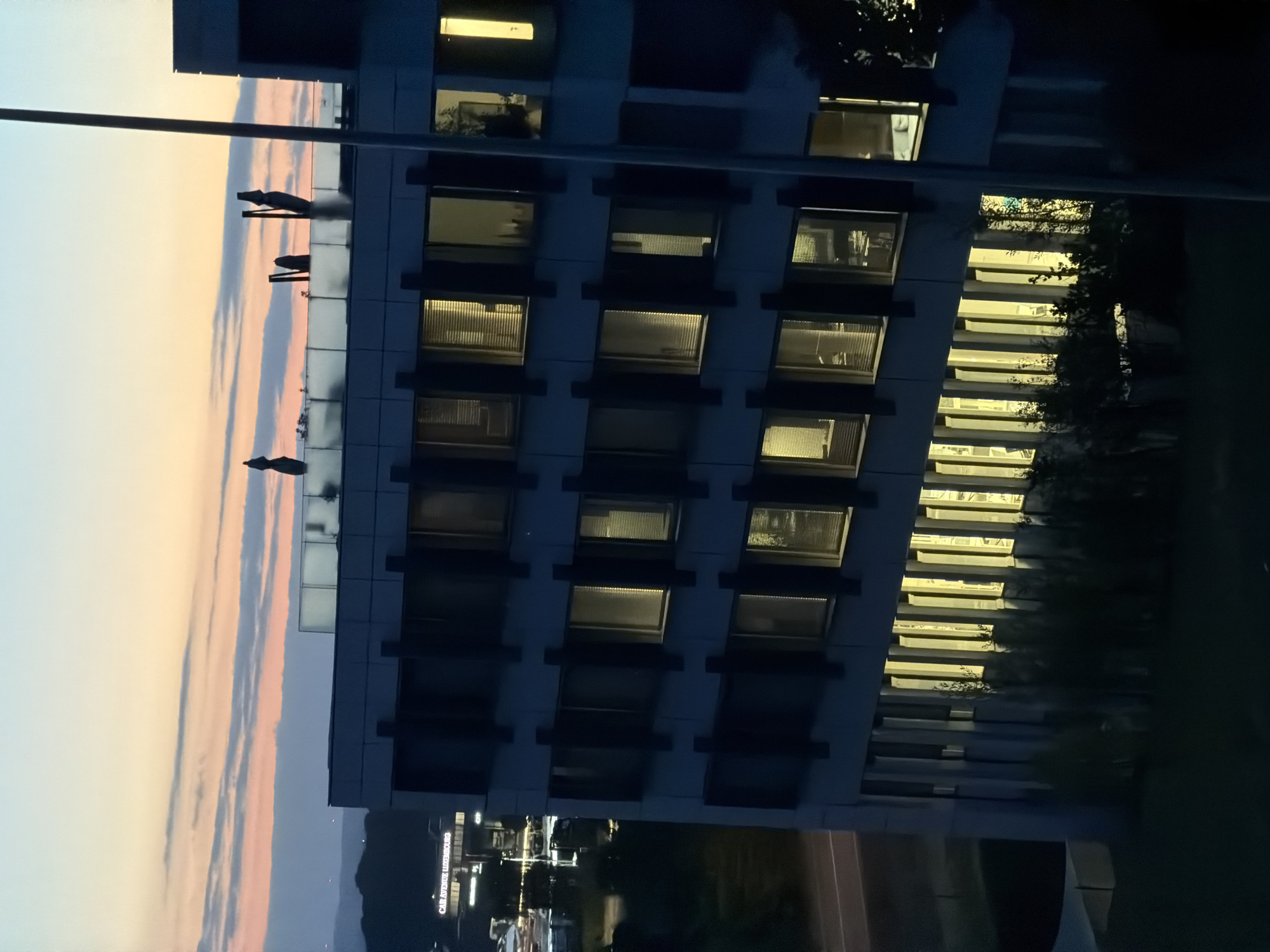 The image shows a modern office building in Leudelange, Luxembourg, at dusk. The sky displays soft hues of orange, pink, and blue from the setting sun, with thin clouds stretching across the horizon. The building's facade is characterized by dark vertical window frames and horizontal bands. Many windows are illuminated from within, casting a warm yellow glow. On the rooftop, several dark, umbrella-like structures and poles are visible, suggesting an outdoor seating or recreational area. In the background, other buildings and lights of a commercial area are faintly discernible. A street or road is visible to the left of the main building, with some greenery in the foreground.