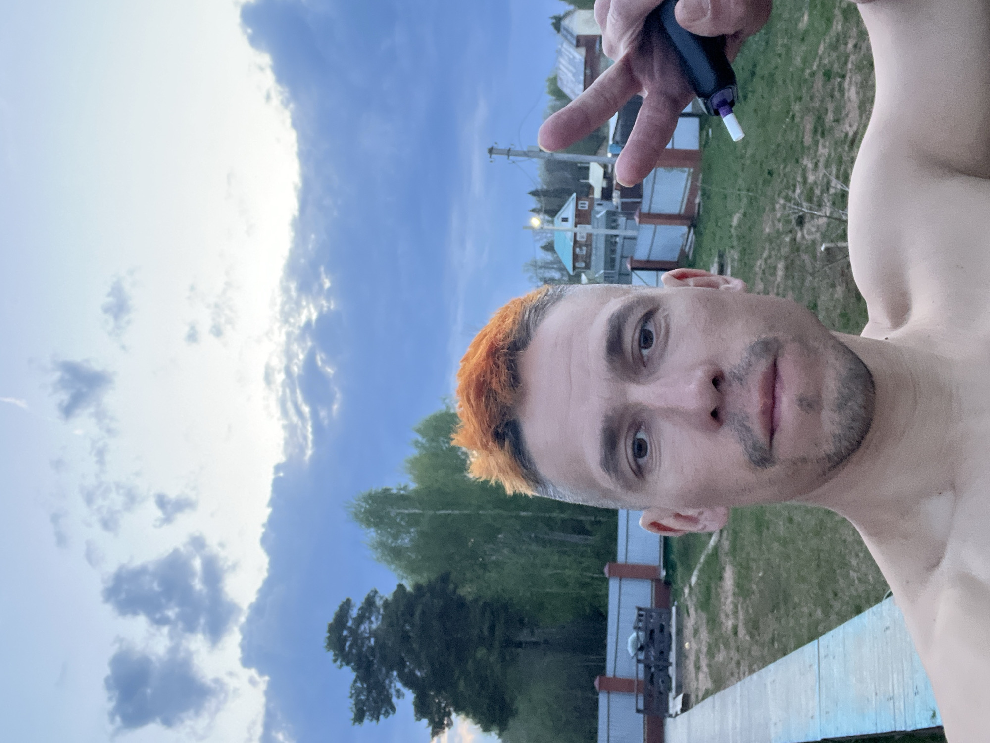 A selfie-style, rotated, medium shot captures a man outdoors, possibly in Chaikovsky, Russia, given the architectural style of houses in the background. The man has orange-dyed hair and stubble. His bare torso is visible. He holds a dark-colored object with a white applicator near his right eye with his left hand while making a peace sign with the same hand. The background features a cloudy blue sky, green trees, and houses with red brick detailing typical of Russian architecture. A white fence and utility poles are also discernible in the scene.