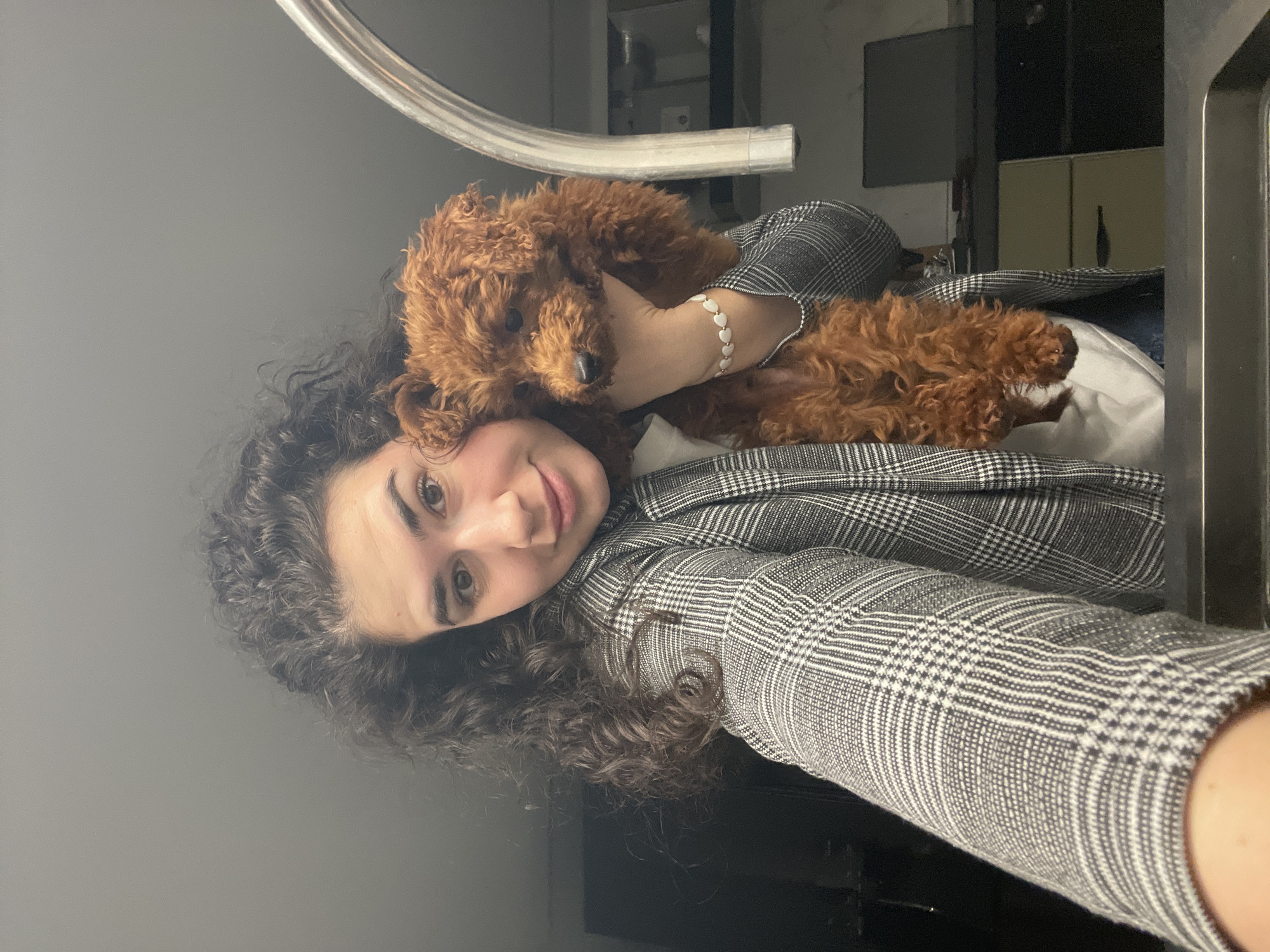 A woman with curly hair wears a plaid blazer and holds a brown poodle. She is smiling. Her left hand is visible and she is wearing a bracelet with heart-shaped charms. The location is indeterminate.