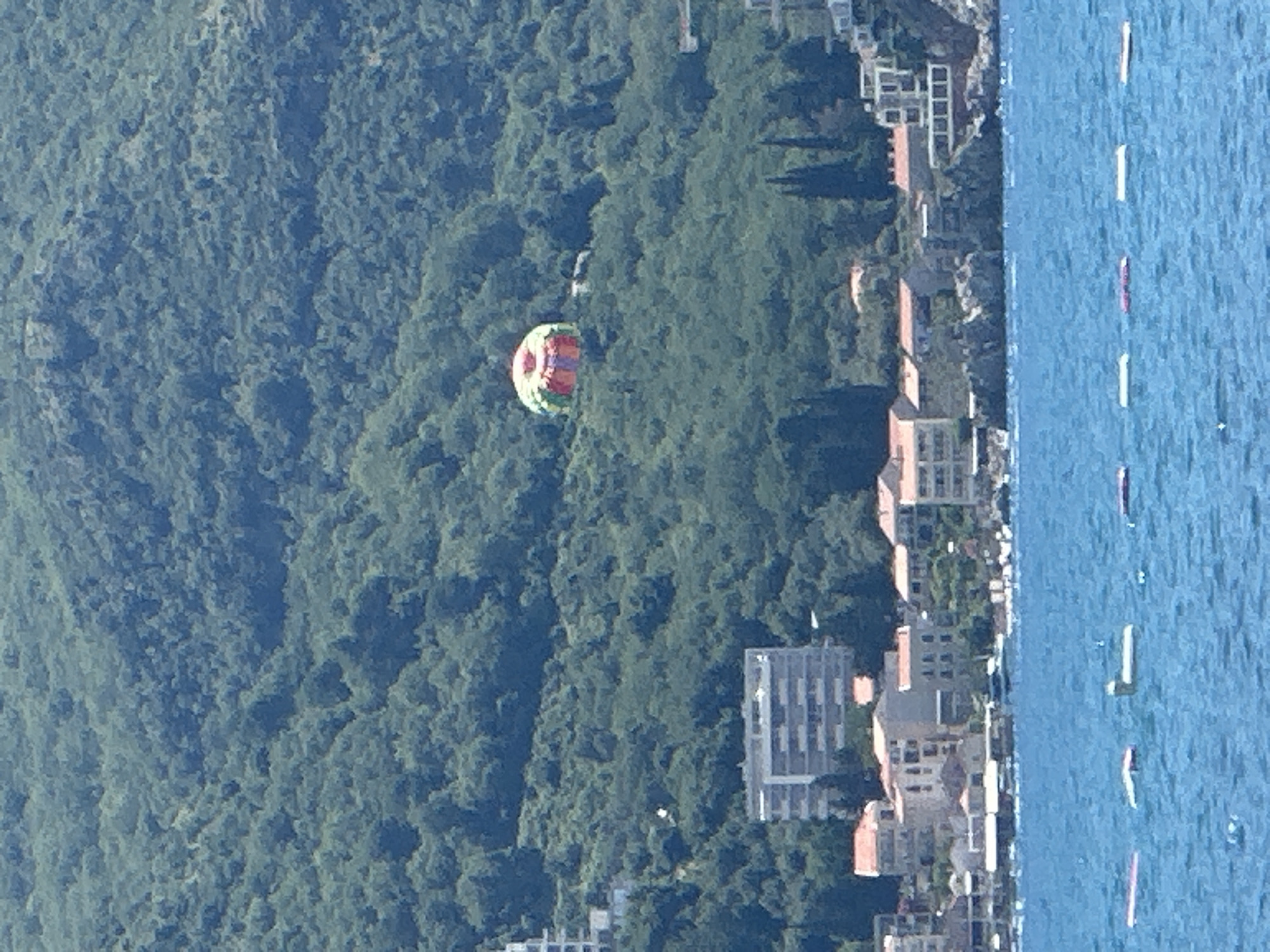 The image captures a coastal scene in Boreti, Montenegro, dominated by a lush, green hillside overlooking a body of blue water. A colorful parachute, likely used for parasailing, is visible against the dense foliage of the hillside. Below the hillside, a cluster of buildings, including hotels and houses with red-tiled roofs, forms a waterfront settlement. The water in the foreground is dotted with a series of buoys, indicating a designated swimming or boating area. The overall impression is one of a serene, sun-drenched coastal resort.