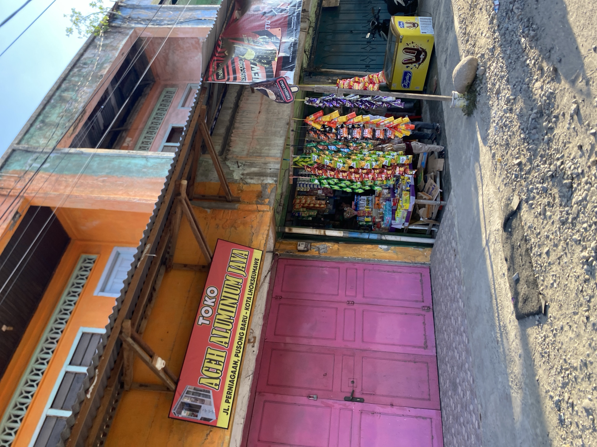 This is an outdoor shot of a small storefront in Lhokseumawe, Indonesia. The store is called "Toko Aceh Aluminium Jaya" and is located on Jalan Perniagaan in Pusong Baru, Lhokseumawe. The storefront has a pink door. Racks of small snack packages hang outside the store. A Joyday-branded ice cream freezer is visible next to the store entrance, and an advertisement banner for "Halococo" ice cream is placed above the snacks. The building next to the store is orange.