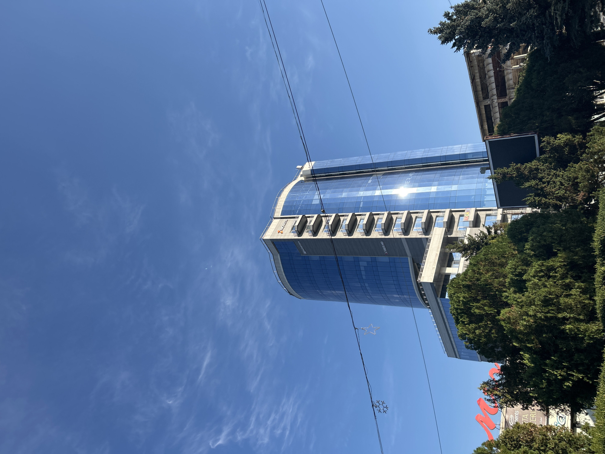 The image shows a modern, curved glass building with multiple balconies, a bright blue sky with wispy clouds, and some trees in the foreground. There are also power lines and some festive star-shaped decorations hanging between them. A red sign with white text, possibly a store name, is visible to the left of the building, peeking through the trees. The building has text on its facade, including "BUSINESSFORM" and "CYBERMD". The sun is reflecting off the glass of the building. The overall scene appears to be an urban environment. The building is identified as the Sun City building in Bălți, Moldova.