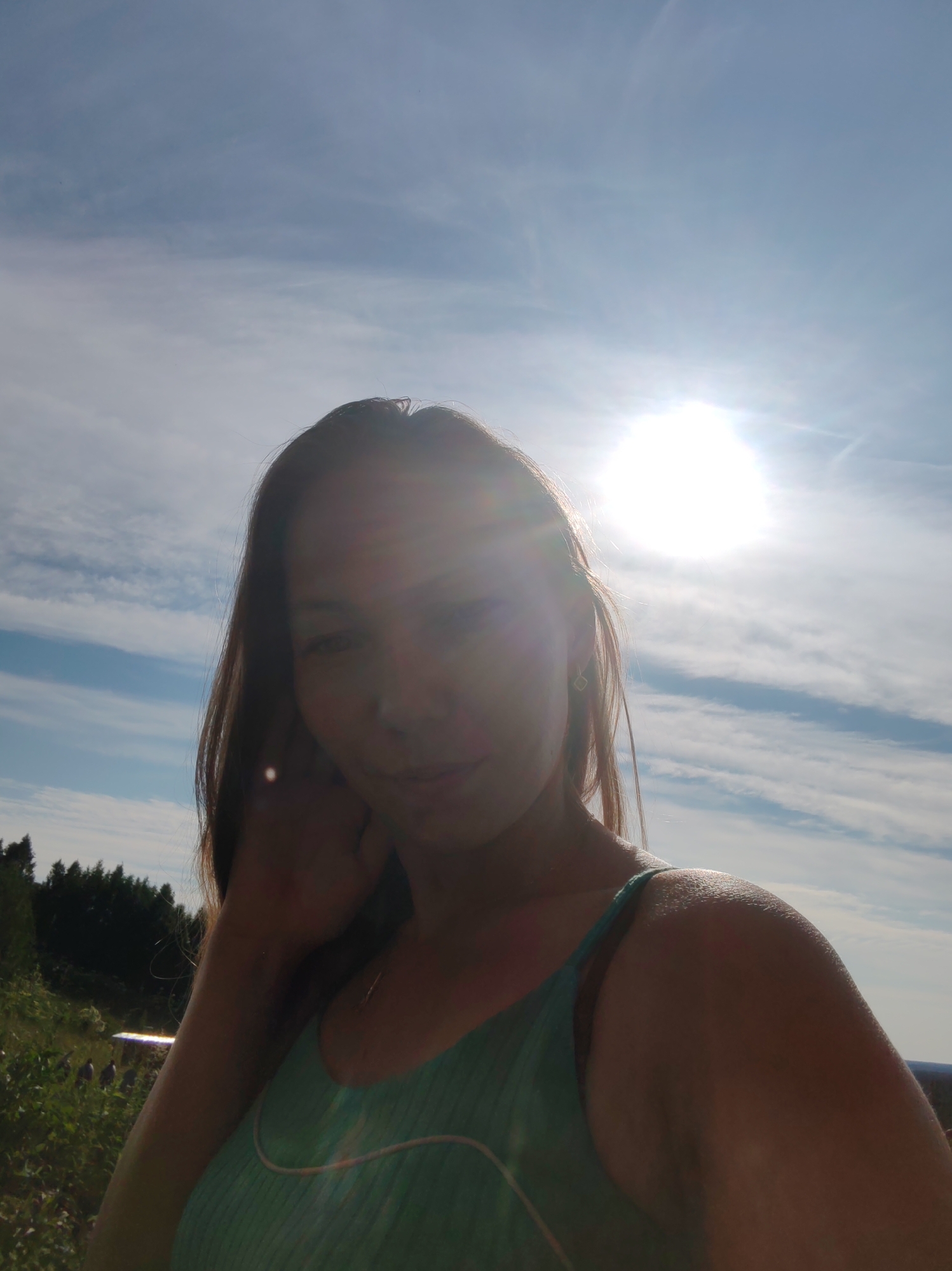 In a sun-drenched outdoor scene, a young woman with long, dark hair takes a selfie against a bright blue sky with scattered clouds. The sun is positioned high in the sky, creating a strong glare that partially obscures her face. She is wearing a teal tank top with thin straps. Her left arm is raised and resting on her head, while her right arm is visible from the shoulder down. In the background, a dense line of dark green trees forms a backdrop against a grassy area. A glimpse of what appears to be a bright, reflective surface, possibly a picnic table or bench, can be seen in the lower left quadrant, with two indistinct figures in the distance. The overall impression is of a casual, summery moment. The location is identified as Chukhlem, Russia.