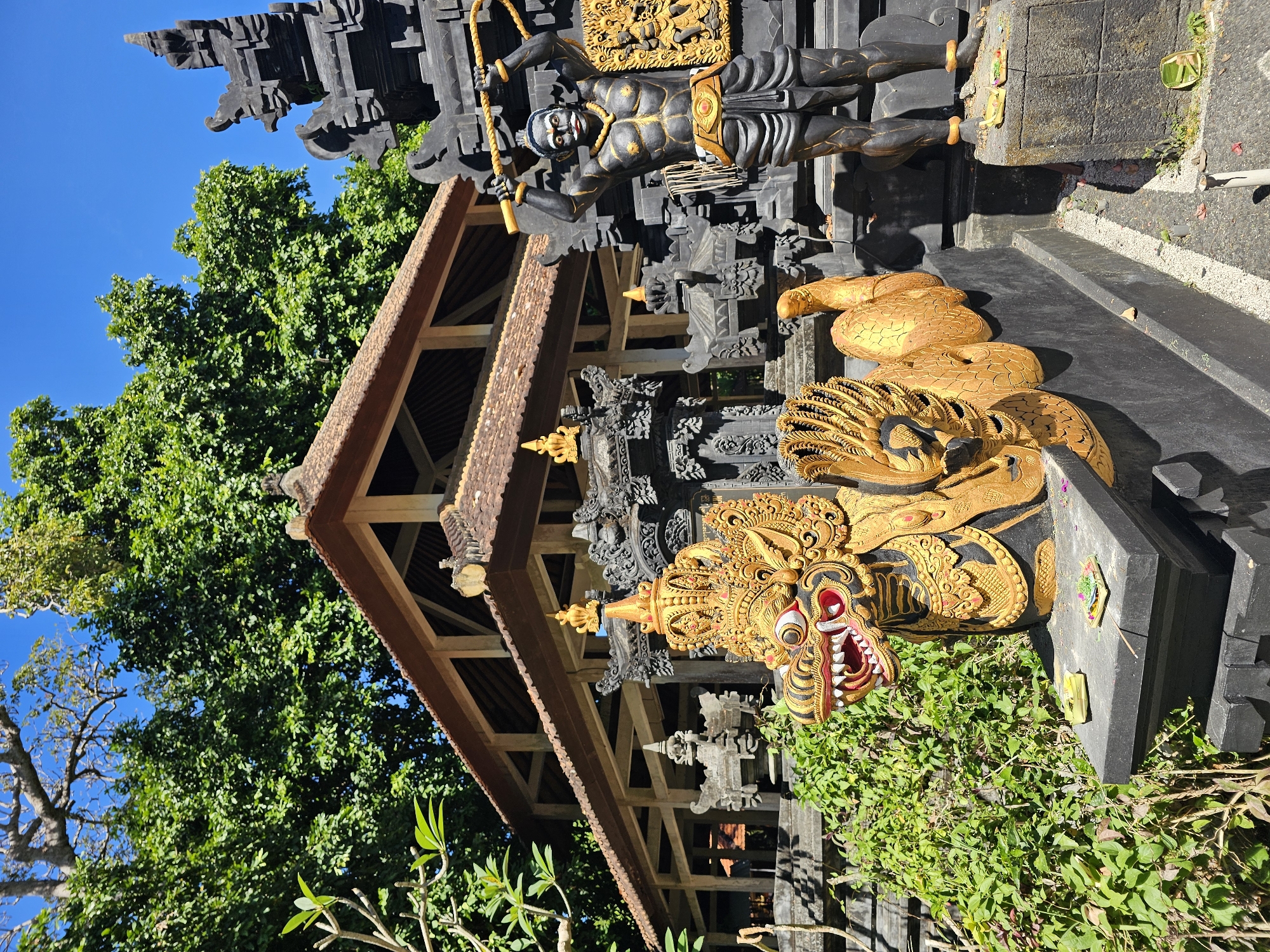 The image shows a traditional Balinese structure in Kuta Selatan, Indonesia. The structure features a multi-tiered roof with brown tiles. The facade is adorned with intricate carvings, including a prominent golden dragon mask and a golden depiction of a snake-like creature. A black statue of a human figure with golden accents stands above. Lush green foliage surrounds the base of the structure, and a clear blue sky is visible in the background.