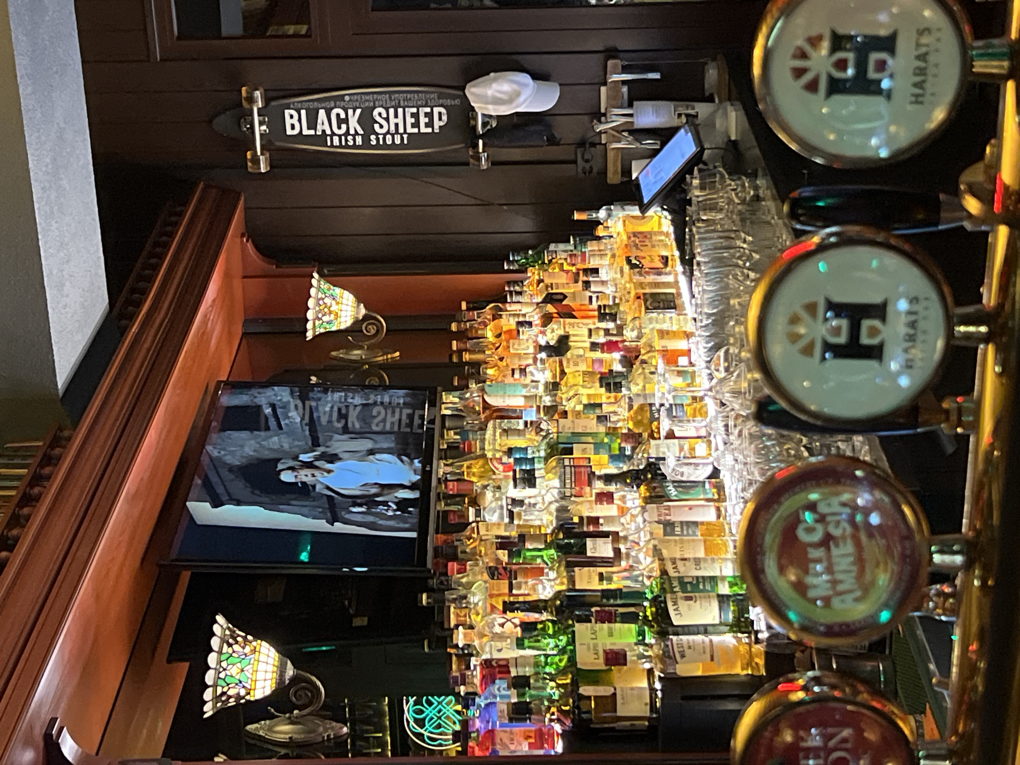 This is a photograph of a bar setting, potentially located in Novosibirsk, Russia, based on the "HARATS" signage, known to be a bar chain present there. The scene features a wooden bar with decorative moldings. A television screen displays an image, possibly promotional, with the words "BLACK SHEEP" mirrored. Above the screen, a skateboard with "BLACK SHEEP IRISH STOUT" printed on it is mounted on the wall, accompanied by a white baseball cap. Below the screen, a Tiffany-style lamp provides ambient lighting, along with a neon cloverleaf sign.
The back wall of the bar is densely packed with bottles, seemingly a display feature. To the right of this bottle display are several beer taps with circular labels; these include "HARATS" and "MELON AMNESIA". The taps are made of brass, suggesting a traditional pub atmosphere. A digital tablet is mounted above the bottle display.
The focus is sharp, with a shallow depth of field highlighting the foreground beer taps, and the general impression is of a dimly lit, bustling bar environment.