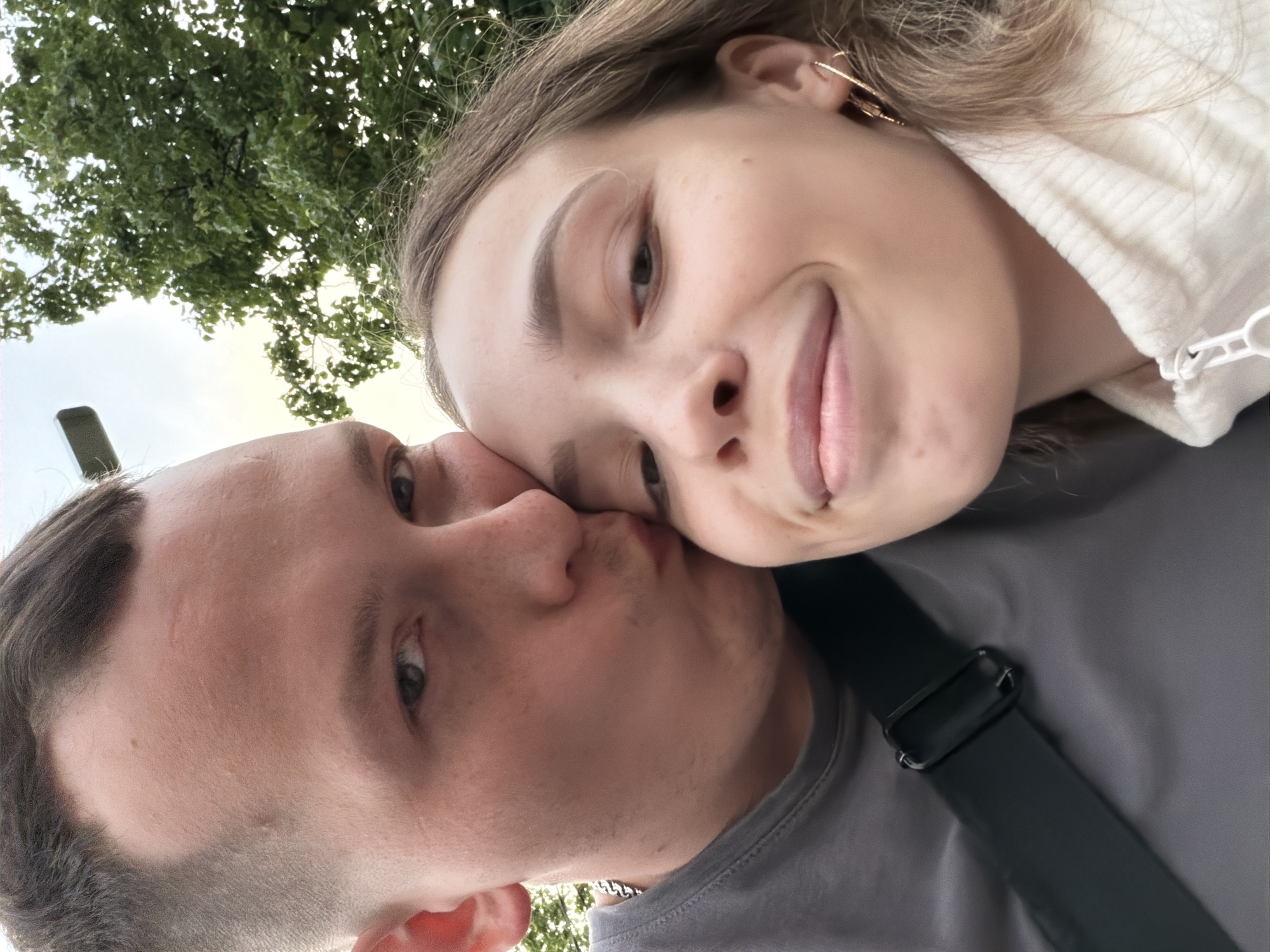 This is a close-up selfie of a young couple outdoors. The man, on the left, is kissing the woman on the cheek. He has short brown hair and is wearing a grey t-shirt. The woman, on the right, is smiling and has long blonde hair. She is wearing a cream-colored turtleneck sweater with a zipper. They are positioned in front of a backdrop of green trees and a cloudy sky. The image appears to have been taken during daylight hours. The location of Chaikovsky, Russia is not visible in the image.