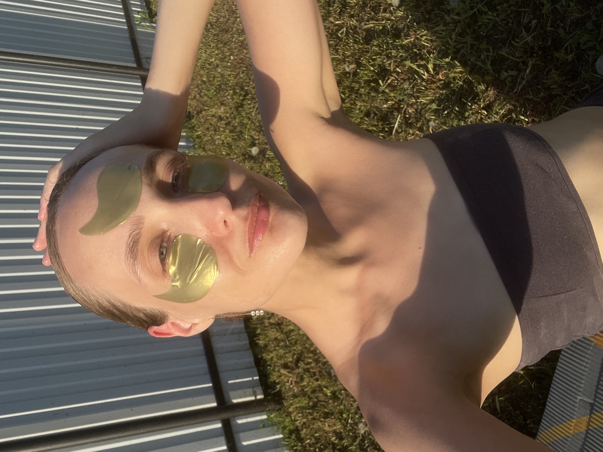 A woman with light skin is lying on her back outdoors. She has gold eye patches on, and is wearing a black strapless top. The woman's right arm is raised over her head, and her left arm is positioned to the side. The background includes grass and a metal structure with horizontal slats. The location is Подшивалово, Russia.