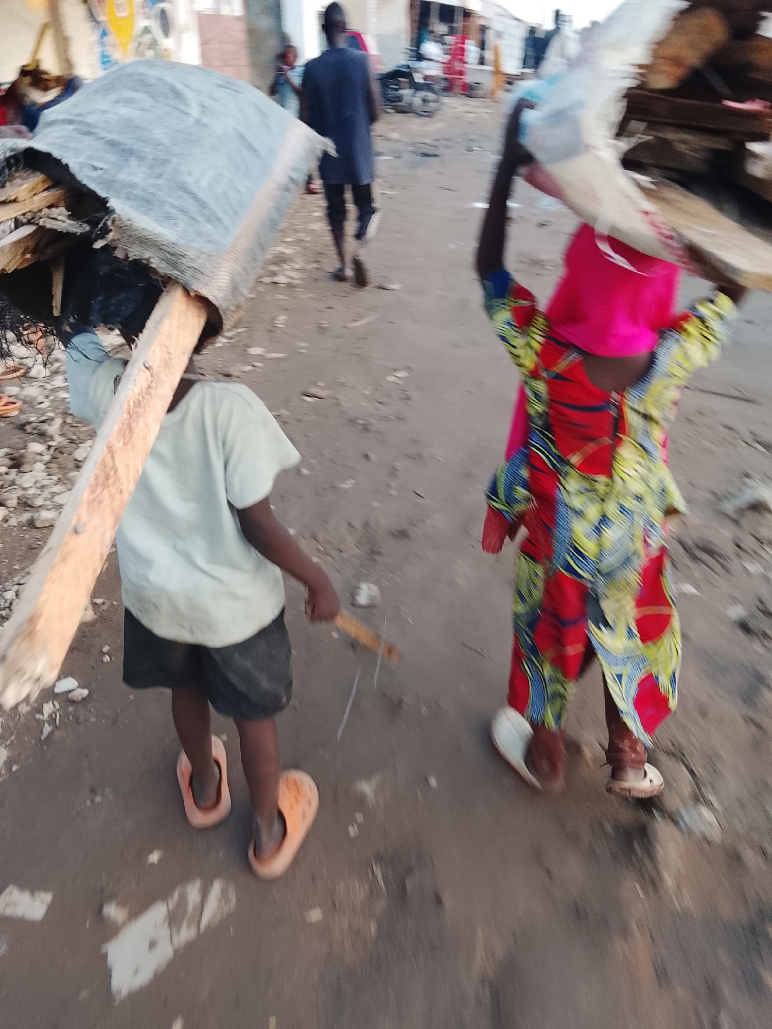 In Abuja, Nigeria, a young child in a light-colored t-shirt and dark shorts is walking, carrying a large, flat piece of debris on their shoulder, supported by a wooden pole. The child is wearing orange clogs. Behind them, another child, wearing a vibrant red, blue, and yellow patterned dress and a pink head covering, is also carrying a load on their head, partially obscured by a plastic bag. They are wearing sandals. A man in a dark blue shirt and dark pants is walking in the background, moving away from the camera. The ground is unpaved and appears to be a dusty or dirt path, with scattered debris and rocks. The overall scene suggests a setting where salvaged materials are being transported.