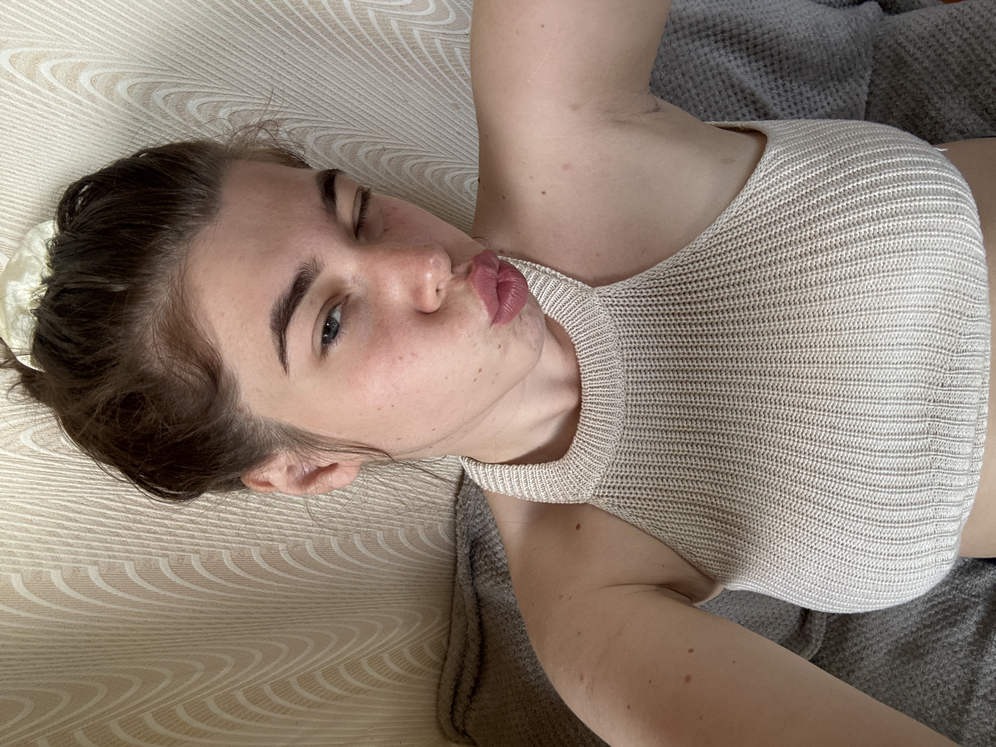 A young woman with her hair pulled up in a bun is shown in a close-up selfie. She is wearing a beige knit halter top. Her left eye is open, and her right eye is closed in a wink. Her lips are pursed as if she is blowing a kiss. The background consists of a beige textured wall with a wavy pattern and a gray blanket. The setting is likely a home interior in Мещёры, Russia.