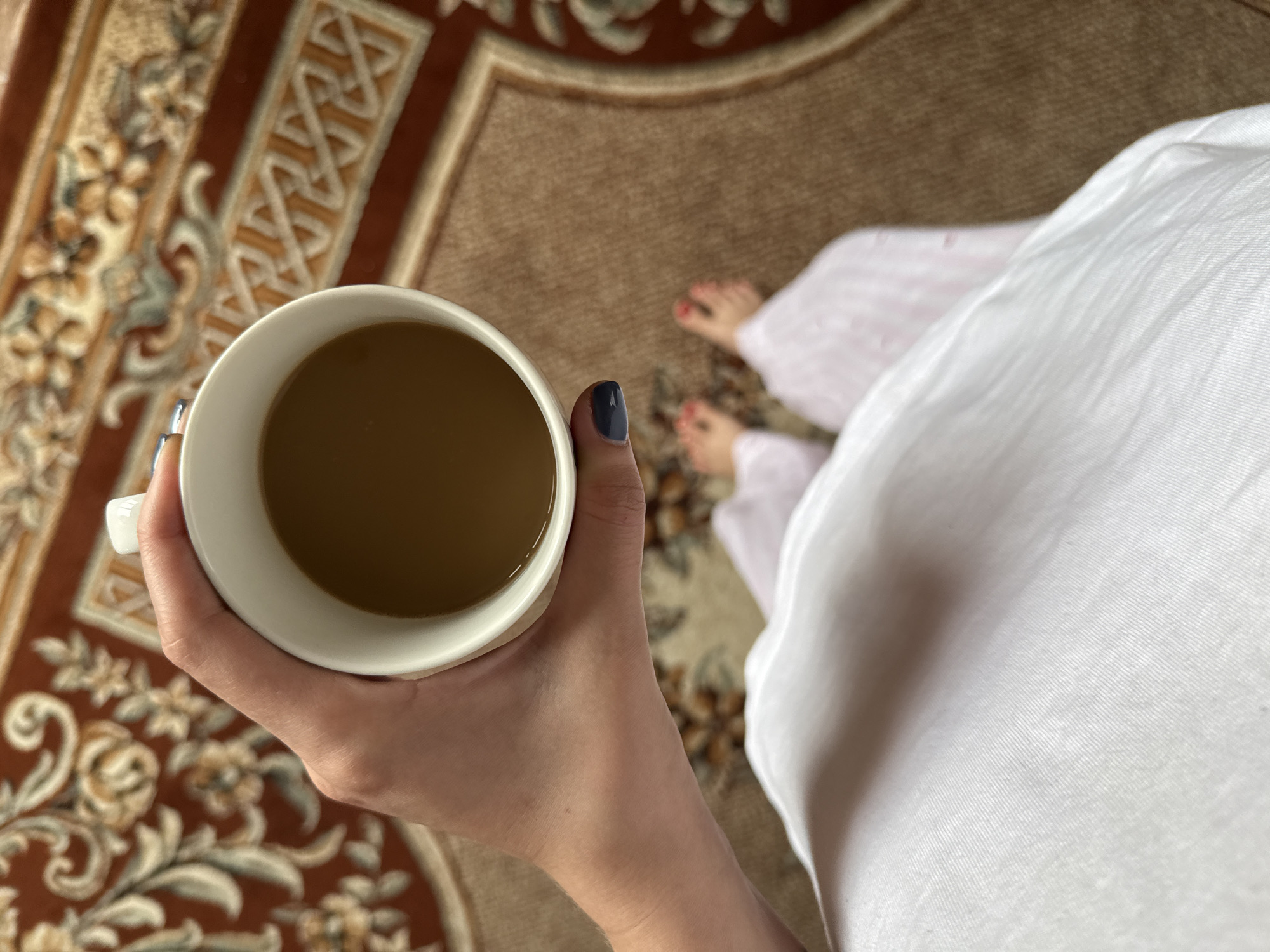A person holds a white mug filled with a brown liquid, possibly coffee or tea. The person's left hand, with a dark-colored nail polish on the thumb, is visible. The individual appears to be wearing a white robe or oversized shirt, with pink-and-white patterned pants visible underneath.
The person is standing on a brown carpet with an elaborate design. The image is taken from a first-person perspective. No information can determine that this is Bely Yar, Russia.
