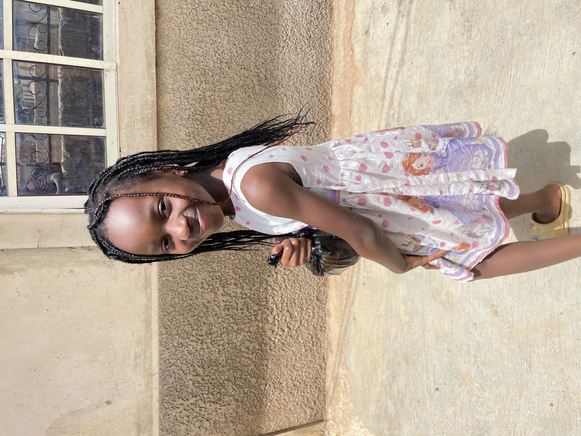 A young African girl stands outdoors near a light-colored wall and window. She has braided hair and wears a white dress with pink polka dots and a purple, cartoon character-themed trim. She holds a black plastic bag in one hand and is smiling. The scene is likely in a sunny location, given the bright light and shadows. Without additional details, conclusively identifying this scene as Jos, Nigeria, is not possible.