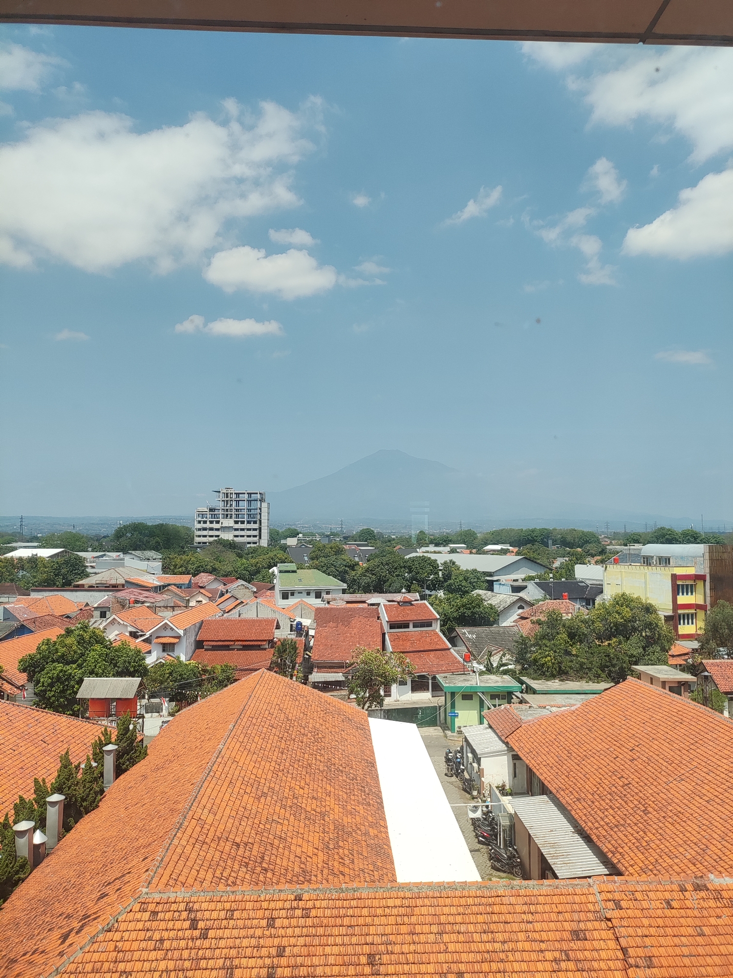 This is an eye-level, outdoor, medium-distance shot of a residential area in Cirebon, Indonesia, with a distant mountain and sky visible. The foreground is dominated by the terracotta tiled roofs of houses. In the mid-ground, more houses with similar tiled roofs are visible, interspersed with green trees and a few modern buildings. One prominent building is a multi-story white structure with blue accents. A large, hazy mountain is visible in the distance against a pale blue sky with scattered white clouds. The scene appears to be captured during the daytime under bright sunlight.