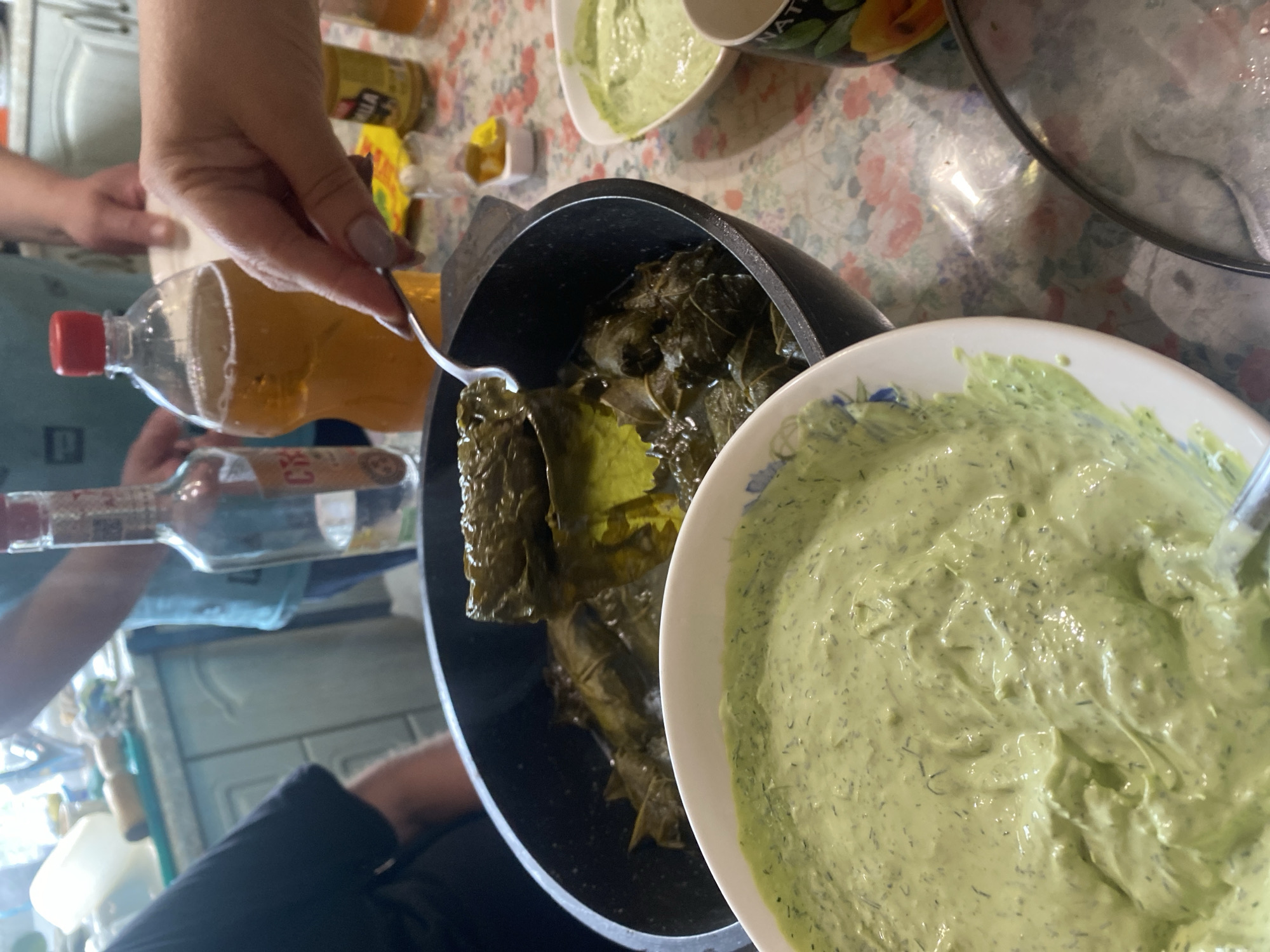 The image depicts a table laden with food, including what appears to be dolmades in a pot and a creamy green dip in a bowl. A person's hand is visible, using a spoon to scoop the dolmades. In the background, bottles of alcohol and a drink are present, along with glimpses of people. The overall setting suggests a meal or gathering. The image is set in the city of Shiryaevo, Russia.