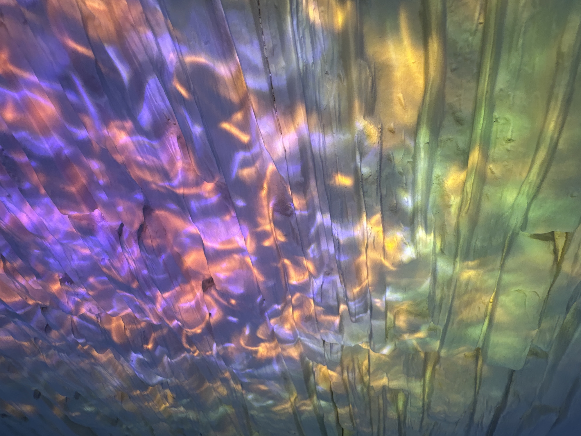 A textured wall, seemingly constructed from translucent, horizontally stacked blocks of ice or a similar crystalline material, is illuminated by dynamic, multi-colored light projections. The surface of the wall exhibits irregular horizontal ridges and undulations, creating a contoured, three-dimensional effect. The projected light forms swirling, organic patterns across the textured surface, reminiscent of water reflections or abstract light art. The coloration of the light transitions across the image: the upper right portion features purples, magentas, and oranges, which gradually shift to yellows and greens in the lower left section. No individuals, additional objects, or specific actions are discernible within the frame, focusing solely on the interaction of light and material. The scene is located in Unknown, Unknown.