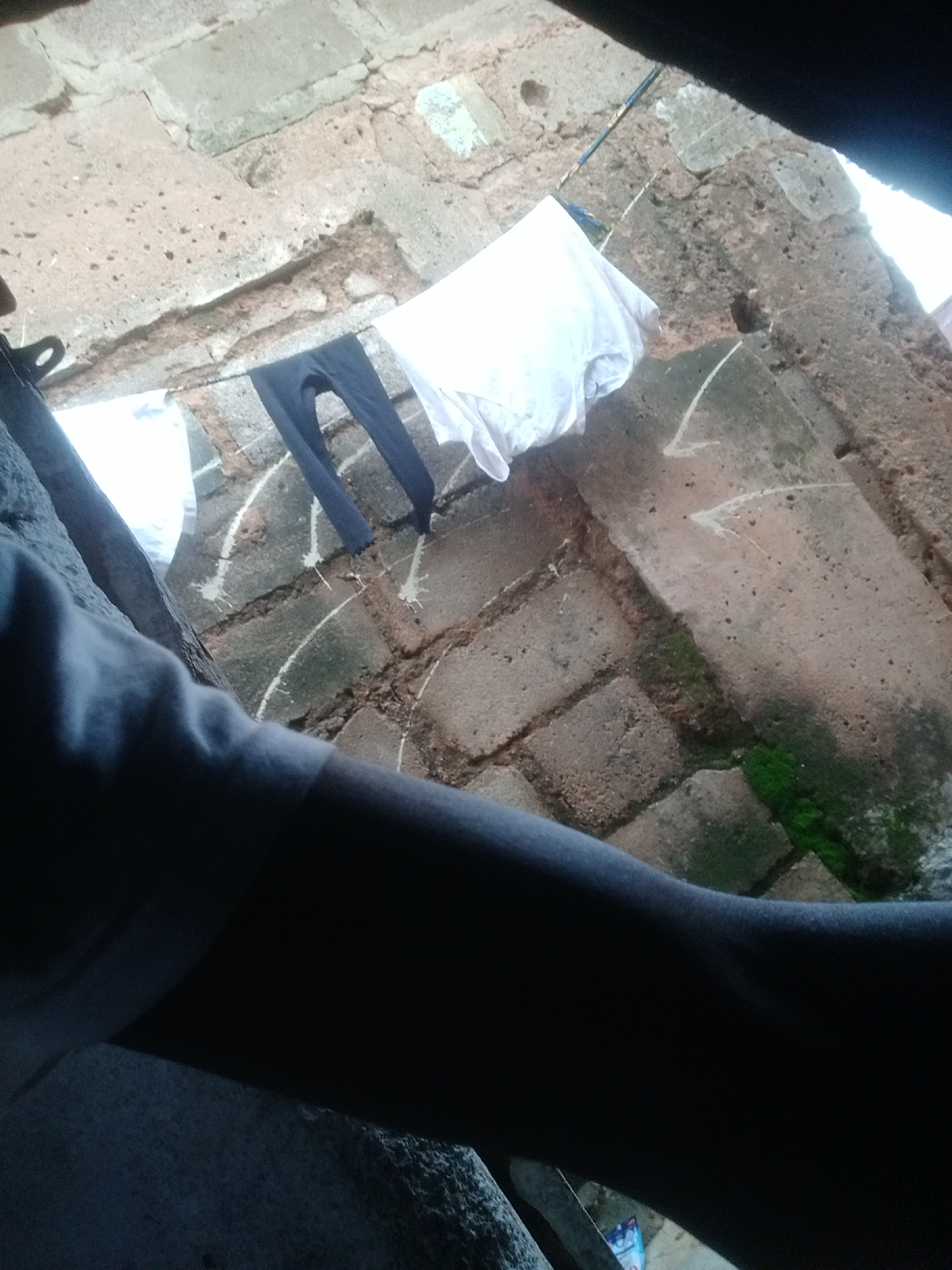 In an outdoor scene, likely in an urban environment, laundry is hanging on a clothesline stretched across a brick wall. Two items of clothing are visible: a pair of dark-colored pants and a white t-shirt. The pants are draped over the line, with the legs hanging down. The t-shirt is a bit further down the line and appears to be a simple white tee. The background consists of a rough, textured brick wall, some of which is covered in moss. The perspective suggests the image was taken from a low angle, possibly looking up at the clothesline and wall. The lighting indicates it is daytime, with some shadows cast on the wall. The overall impression is one of a domestic setting where laundry is being air-dried.