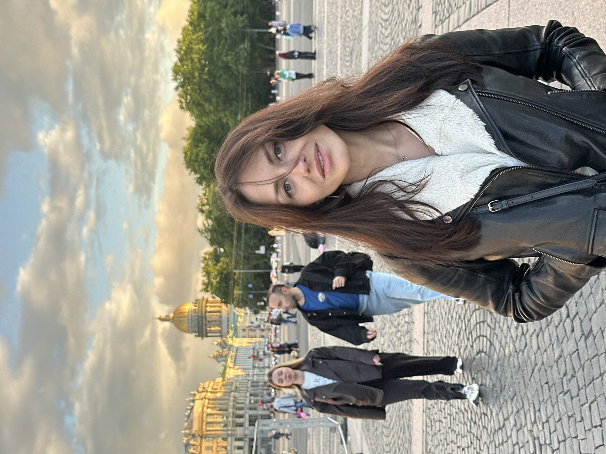 The image depicts a group of people in an outdoor urban setting, likely Saint Petersburg, Russia, given the architectural style of the buildings and the presence of a golden dome, which resembles St. Isaac's Cathedral. The sky is filled with dramatic clouds, suggesting either early morning or late afternoon.
The main subject is a young woman in the foreground, captured in a selfie-style shot, with her face turned towards the camera. She has long, dark brown hair and is wearing a black leather jacket with a white furry lining and a silver necklace. Her expression is neutral, with a slight smile.
Behind her, other individuals are visible. One man in a blue t-shirt and black jacket is walking away from the camera. Further in the background, other figures are seen, some appearing to be tourists or pedestrians. The ground is paved with cobblestones. The overall atmosphere appears relaxed, possibly during a leisurely stroll or outing. The lighting suggests it is either sunrise or sunset, casting a warm glow on the scene. No visible text is present in the image.