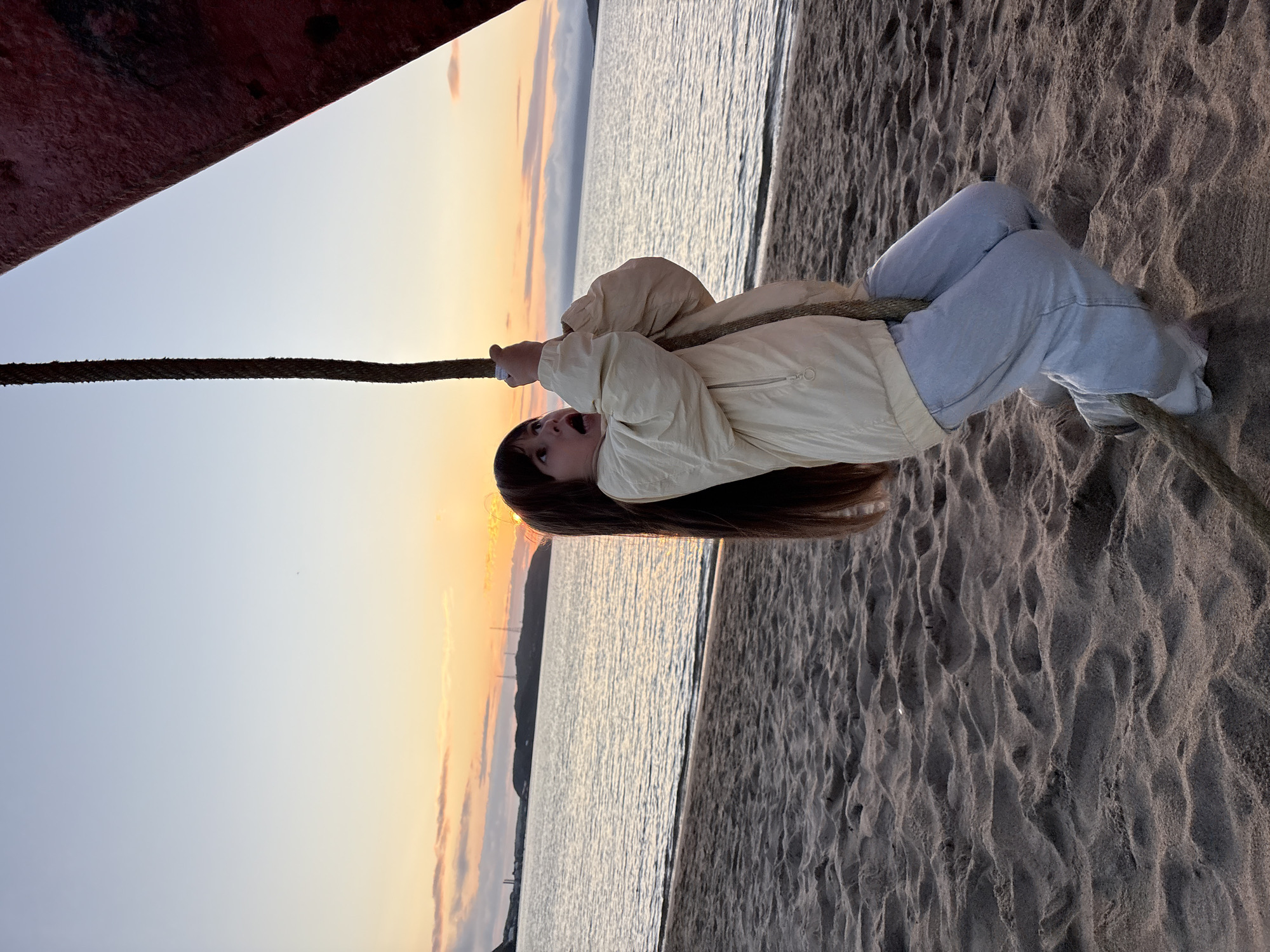 A young girl with long brown hair is holding onto a thick rope, appearing to swing or climb. She is wearing a light yellow jacket and light blue jeans. Her mouth is open in surprise or excitement. The rope is attached to a large red object on the right side of the frame, possibly part of a structure or vessel. The girl is standing on a sandy beach. In the background, a calm body of water stretches to the horizon, with a silhouetted landmass and some structures visible in the distance. The sky displays a soft sunset with hues of orange, yellow, and pink. The scene is set at sunset on a beach in Teriberka, Russia, a coastal village on the Kola Peninsula known for its dramatic Arctic landscapes and proximity to the Barents Sea.