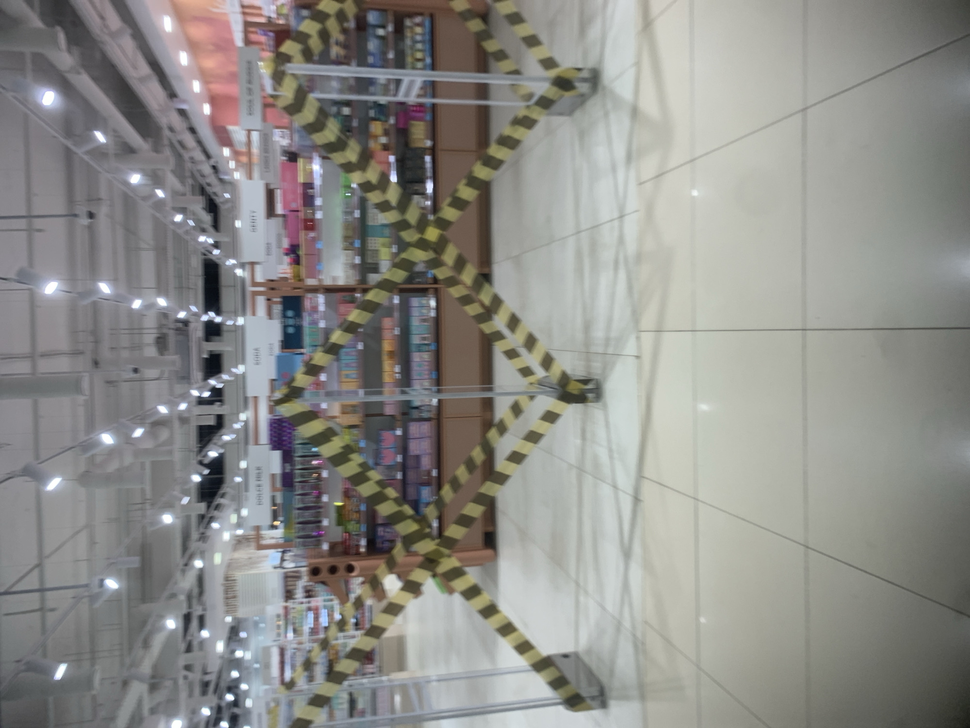 The image displays the interior of a brightly lit store aisle, viewed from a slightly elevated perspective. The light-colored floor is composed of large square tiles with dark grout lines.
A prominent yellow and black striped accordion-style barrier stretches across the aisle, effectively blocking access. This barrier casts strong, distinct shadows onto the tiled floor to its right. To the left of the barrier, several shelves are visible, stocked with numerous products that appear brightly colored, suggesting items such as books, stationery, or cosmetics. The shelving units are made of what appears to be wood or a similar material, with metal supports.
Above the shelves, multiple white rectangular signs are suspended from the ceiling, indicating product categories or sections. Visible text on these signs includes: "BOOK OF AUDI..." (partially obscured), "LARGE BERRIES", "BEAUTY", "ROGA", and "HOLEY MILLE" (partially visible). These labels suggest a diverse retail environment, possibly a bookstore with other sections, a large department store, or a beauty supply store.
The white ceiling features numerous rectangular artificial lights arranged in rows, providing extensive illumination to the space. White structural elements, likely beams or conduits, are also present on the ceiling.
No individuals are visible in the image. The presence of the barrier indicates that this specific aisle or section of the store is temporarily closed, undergoing maintenance, or otherwise restricted. The setting is an indoor commercial space in Izhevsk, Russia.