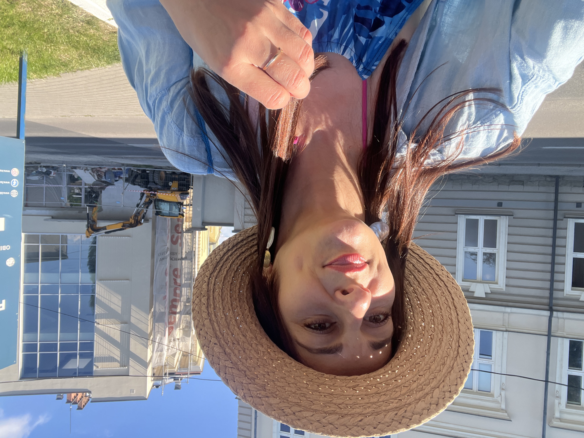 A woman wearing a straw hat and a light blue shirt is taking a selfie outdoors. She has long brown hair, brown eyes, and is wearing earrings and a ring. She is smiling and looking at the camera. In the background, there is a building under construction with scaffolding and a yellow excavator. The scene appears to be in a city.