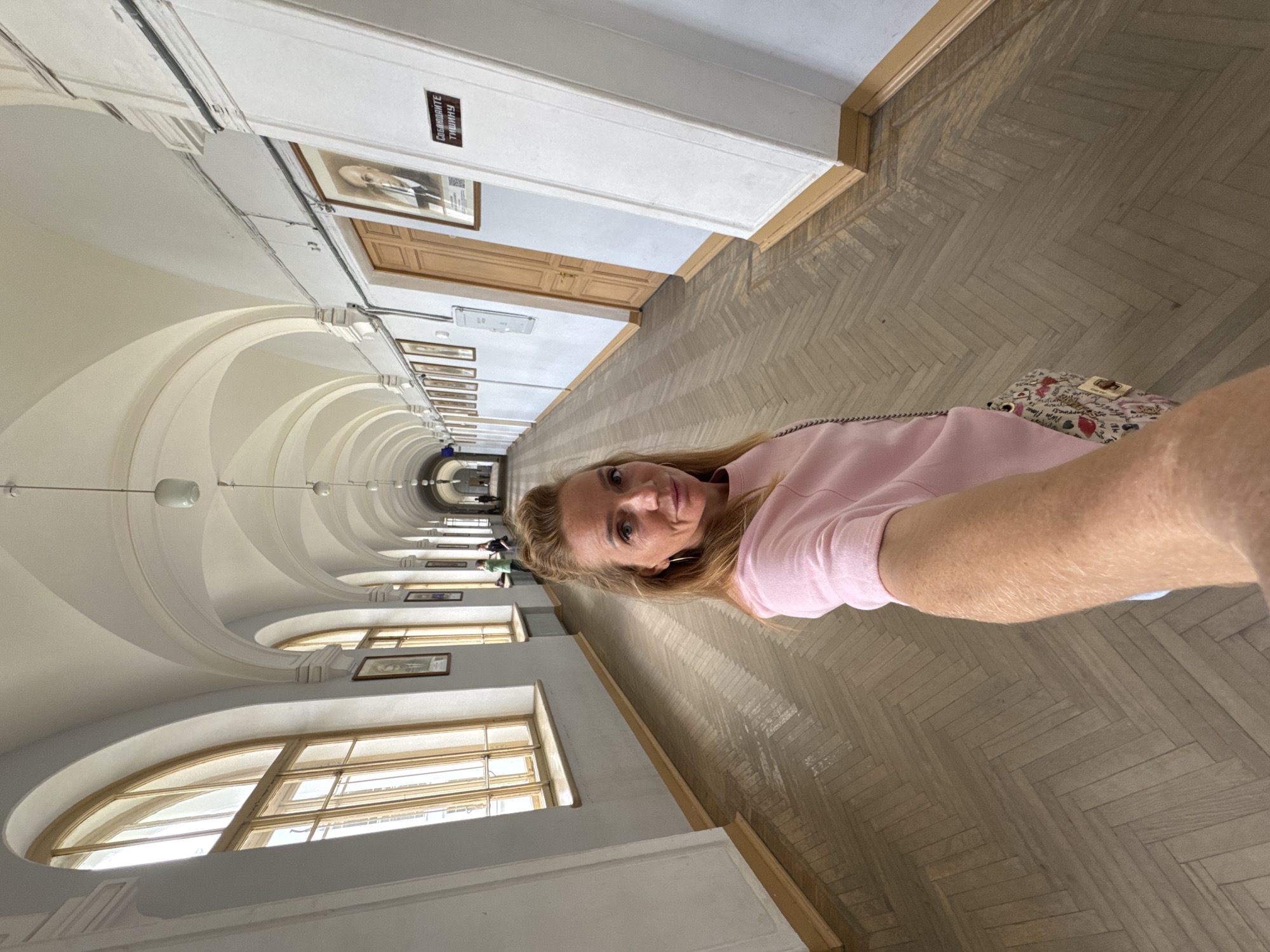 The image depicts a woman taking a selfie in a long, arched hallway. The hallway has a high, vaulted ceiling with multiple arched windows on the left side, letting in natural light. The floor is made of herringbone-patterned hardwood. Along the right wall, there are several framed portraits and a wooden door. A sign with Russian text is visible on the wall. In the distance, other people can be seen walking down the hallway. The woman is wearing a pink sweatshirt and carrying a small patterned bag. This hallway is likely located within a historical building in Saint Petersburg, Russia, possibly a university or administrative building, given the architecture and the portraits displayed.