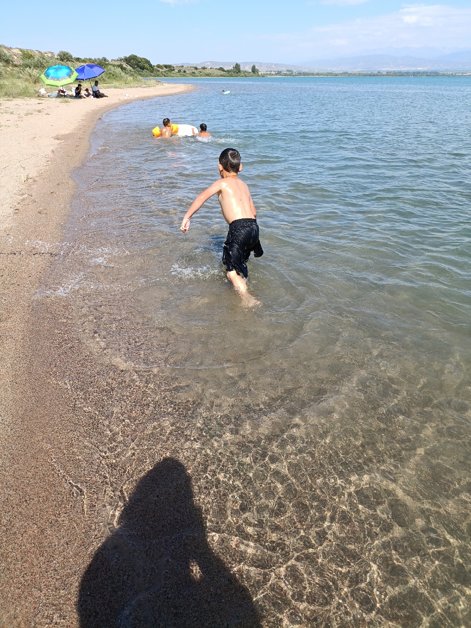 In the city of Kirpichny, Kyrgyzstan, a boy is walking in shallow water near a sandy beach. The boy is wearing black shorts and is facing away from the camera, with his arms extended slightly. Several other individuals are visible in the background, some sitting under umbrellas on the shore and others in the water with inflatable toys. The water is clear, allowing the sandy bottom to be seen. The environment is a lake or a large body of water, with a shoreline that transitions from sand to a grassy embankment. Distant hills and a clear blue sky complete the scene.