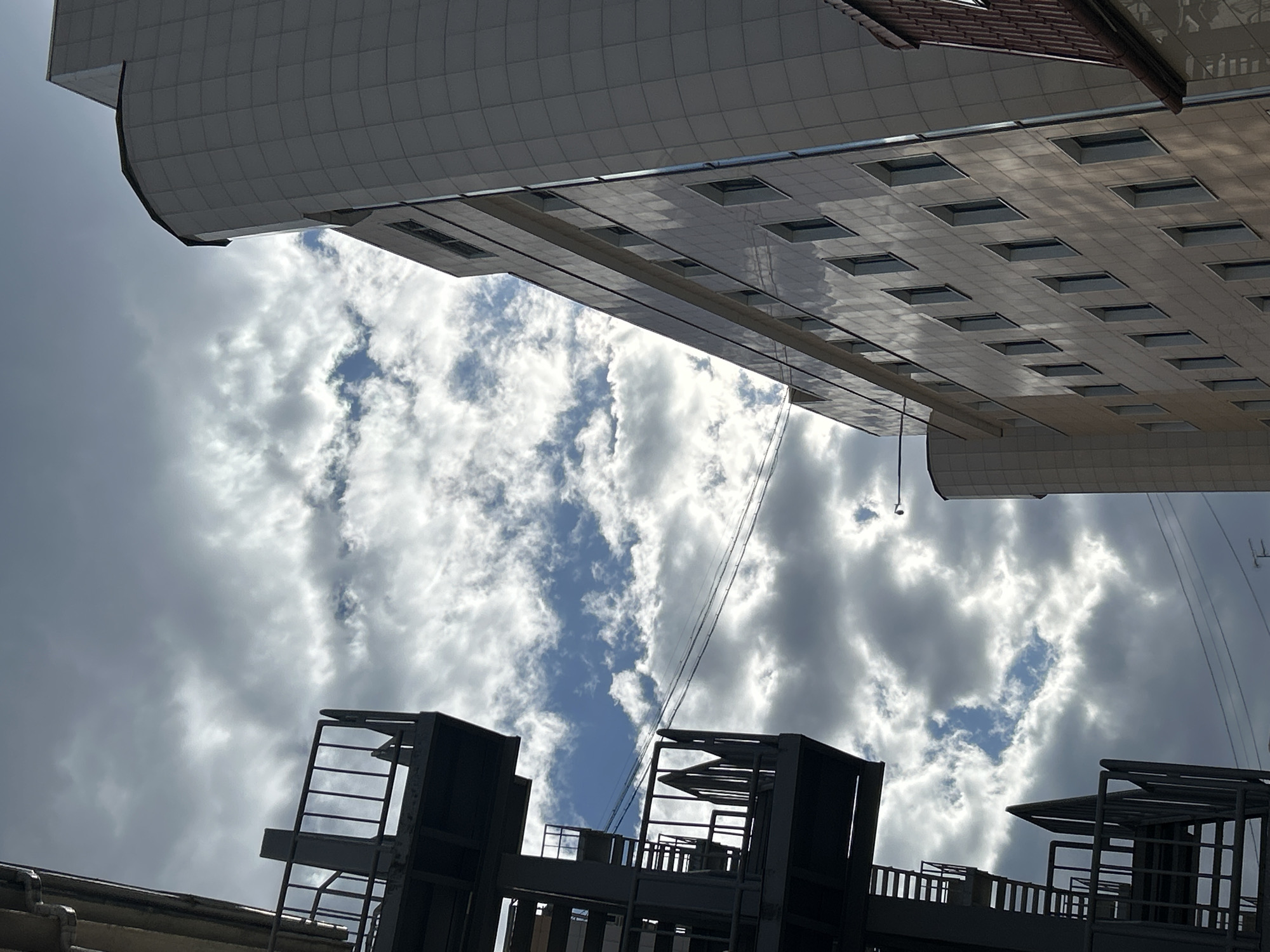This upward-angled photograph captures modern architectural elements against a dynamic, partially cloudy sky in Moscow, Russia. The image is composed with two distinct building sections framing the sky.
In the upper and right portions of the frame, a large, light-grey building with a geometric, tiled or paneled facade is visible. Its underside or an extended overhang features numerous regularly spaced square openings, which appear to be windows or skylights, reflecting the sky's light. A small section of a reddish-brown tiled roof is also visible at the top right, indicating another part of the building.
The central and largest part of the image is filled with a bright, partially cloudy sky. White cumulus clouds, brightly lit by the sun, intermingle with darker grey cloud formations, revealing patches of clear blue sky, especially towards the middle. Several thin, dark power lines or cables diagonally traverse the sky in the middle-right section.
In the lower part of the image, a dark, industrial-looking structure or section of another building occupies the foreground. This structure features prominent railings, grates, and an open framework, suggesting a rooftop installation, a walkway, or a complex architectural feature.
The scene takes place during the daytime under bright, diffused sunlight. No people are visible, and there is no discernible text in the image. The unique perspective emphasizes the architectural design and the interplay between man-made structures and the natural sky.