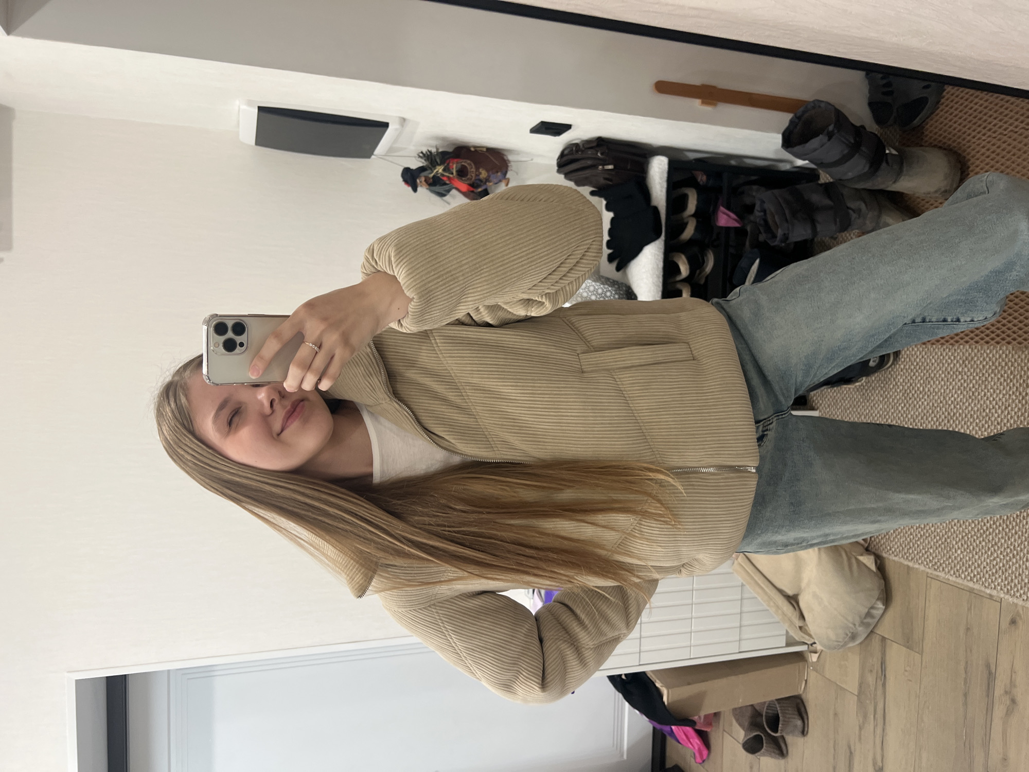 A young woman with long, straight light brown hair is captured in a mirror selfie, smiling softly with her eyes gently squinting. She holds a silver smartphone with a clear case in front of her face, and a ring is visible on her finger. She is wearing a light beige or tan ribbed jacket with puffed sleeves and a zipper, paired with light blue denim jeans and a white top underneath.
The setting appears to be an indoor room, likely a hallway or bedroom in Votkinsk, Russia. The background, reflected in the mirror, shows plain white walls, a light-colored wooden floor, and a section of light brown textured carpet. To the right of the woman (left in the reflection), a shoe storage unit or shelf holds various items, including dark boots, other shoes, and black gloves. Above this, a brown bag with decorative elements and possibly a hat are visible. A black rectangular object, possibly a speaker or air vent, is mounted on the wall. A white door frame is also visible to the woman's left. The overall impression is a casual, domestic scene.
