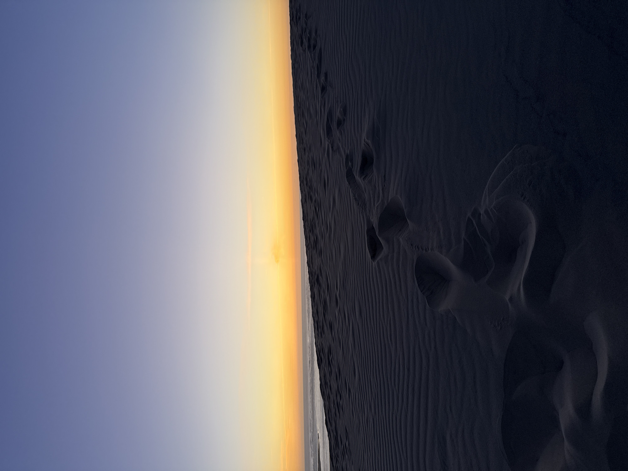 The image depicts a dark sand dune landscape at dusk, likely in the Dune du Pilat, located in La Teste-de-Buch, France. The sky transitions from a deep blue at the top to a soft yellow and orange band near the horizon where it meets the ocean. Footprints are visible in the rippled sand, leading towards the sea. The foreground is dominated by the dark sand, with subtle undulations and shadows. The overall impression is one of solitude and the passage of time, marked by the footprints.