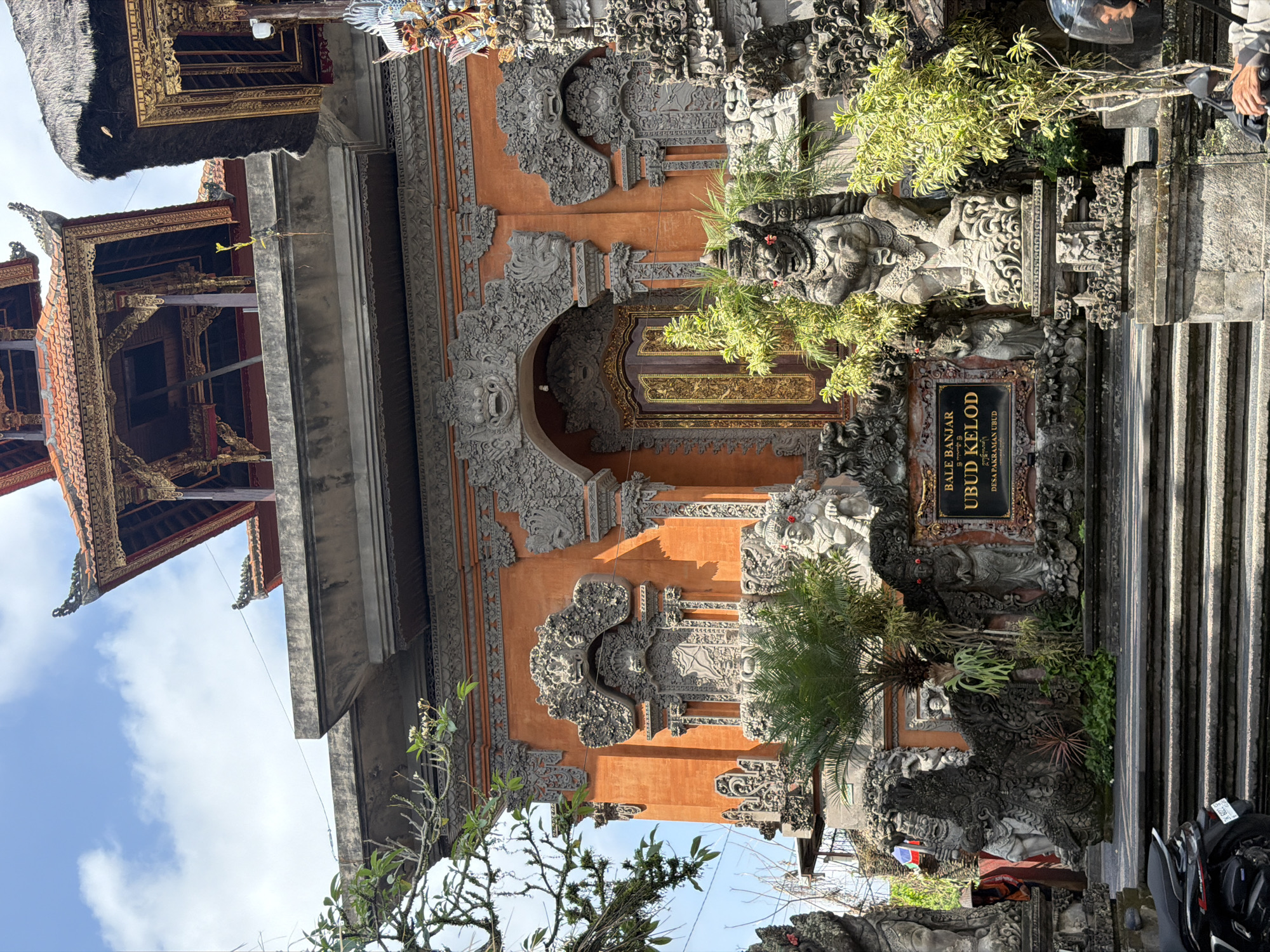 The image shows the entrance to the Bale Banjar Ubud Kelod, a cultural center in Ubud District, Indonesia. The entrance features traditional Balinese architecture with ornate stone carvings and a prominent orange facade. Several stone statues of mythical creatures and figures guard the entrance. A signpost in front of the building reads "BALE BANJAR UBUD KELOD DESA PAKRAMAN UBUD." Lush greenery, including palm trees and smaller plants, surrounds the statues and the building. In the foreground, a motorcycle is parked on the steps leading to the entrance, and a person wearing a helmet is visible on the motorcycle. The sky above is partly cloudy.